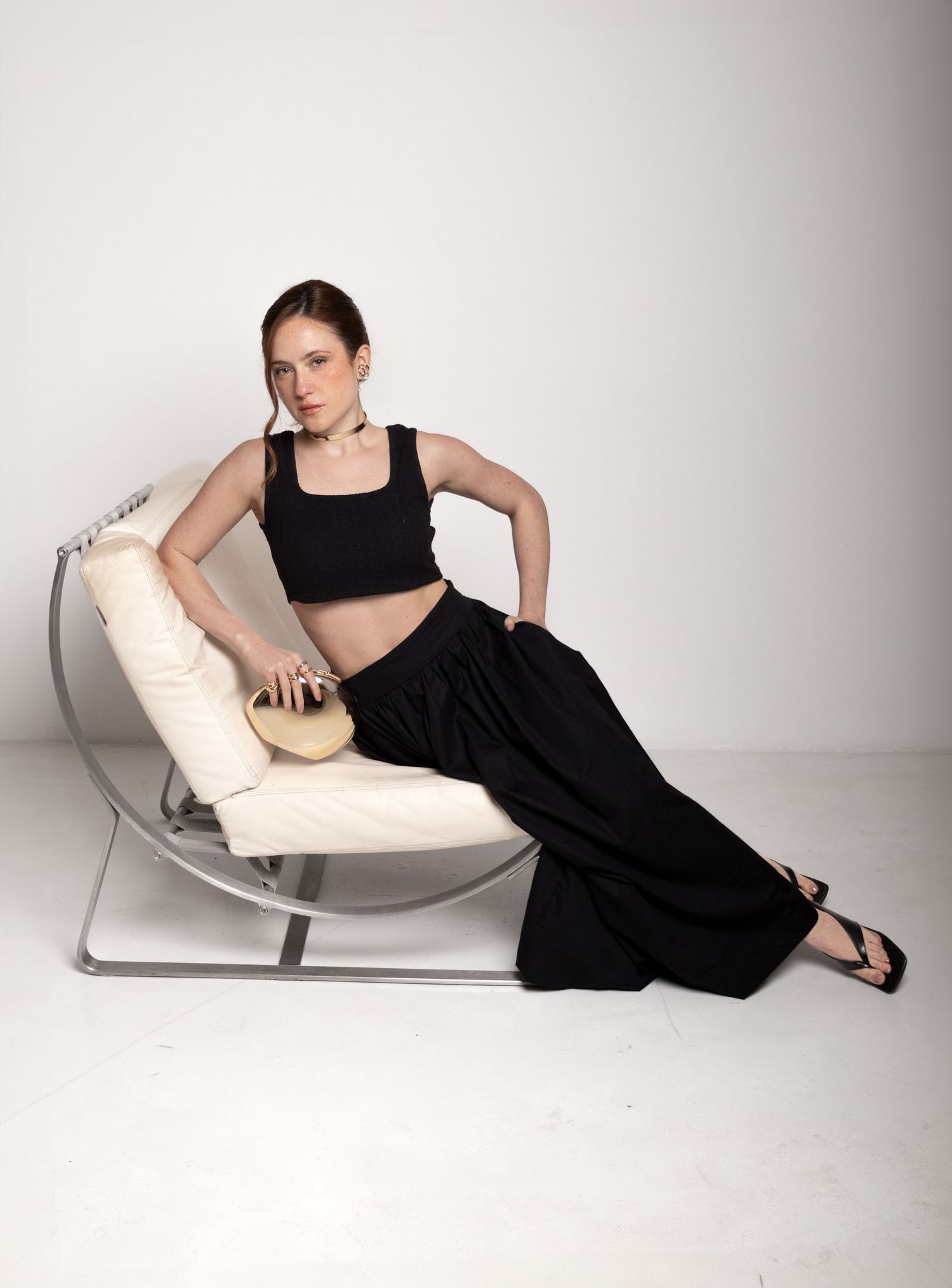 Model lounges on a curved cream chair in a black crop top and wide black skirt, holding a small gold clutch.