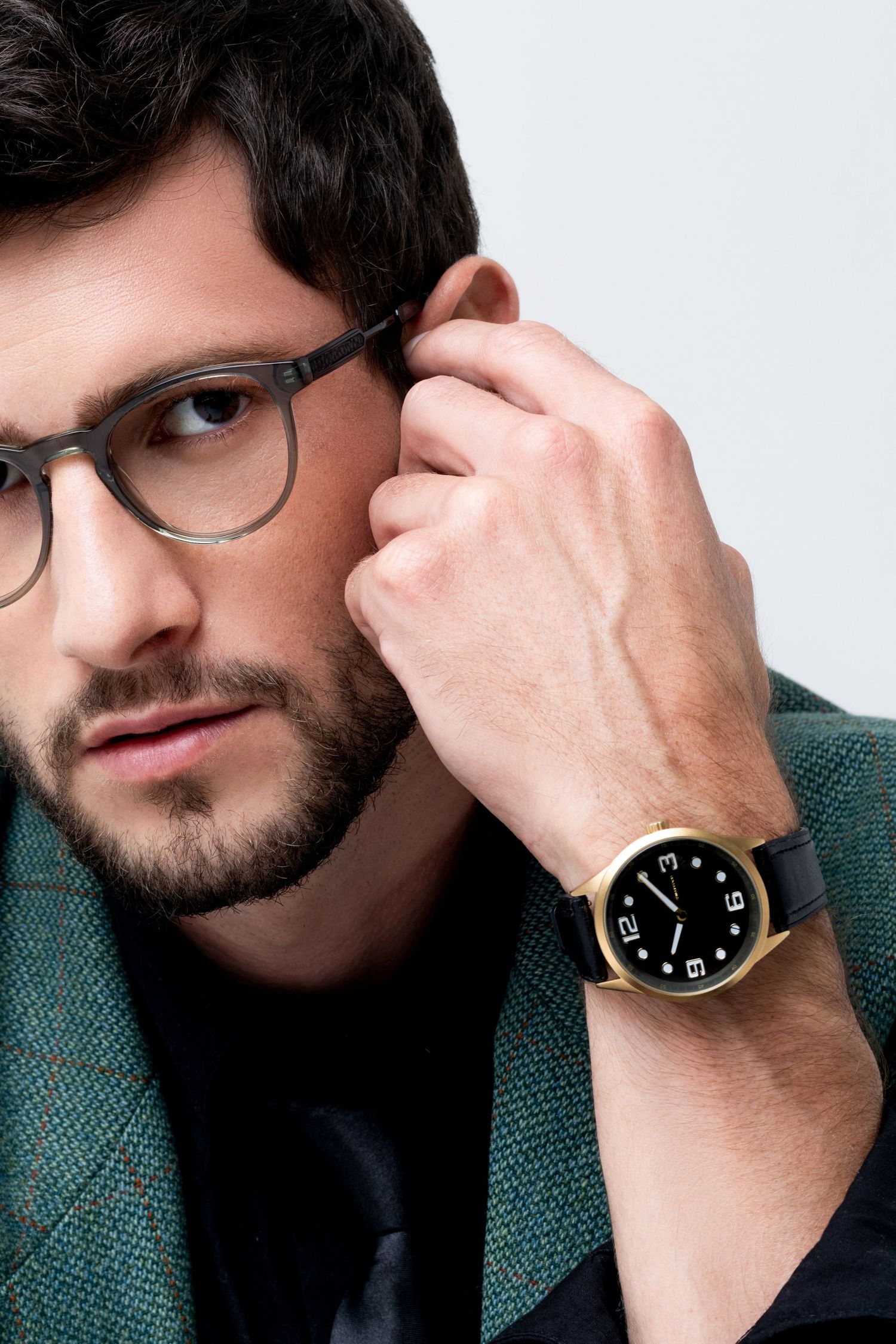 Bearded man in gray-framed glasses touches his ear, wearing a gold-cased watch with black dial and black leather strap.