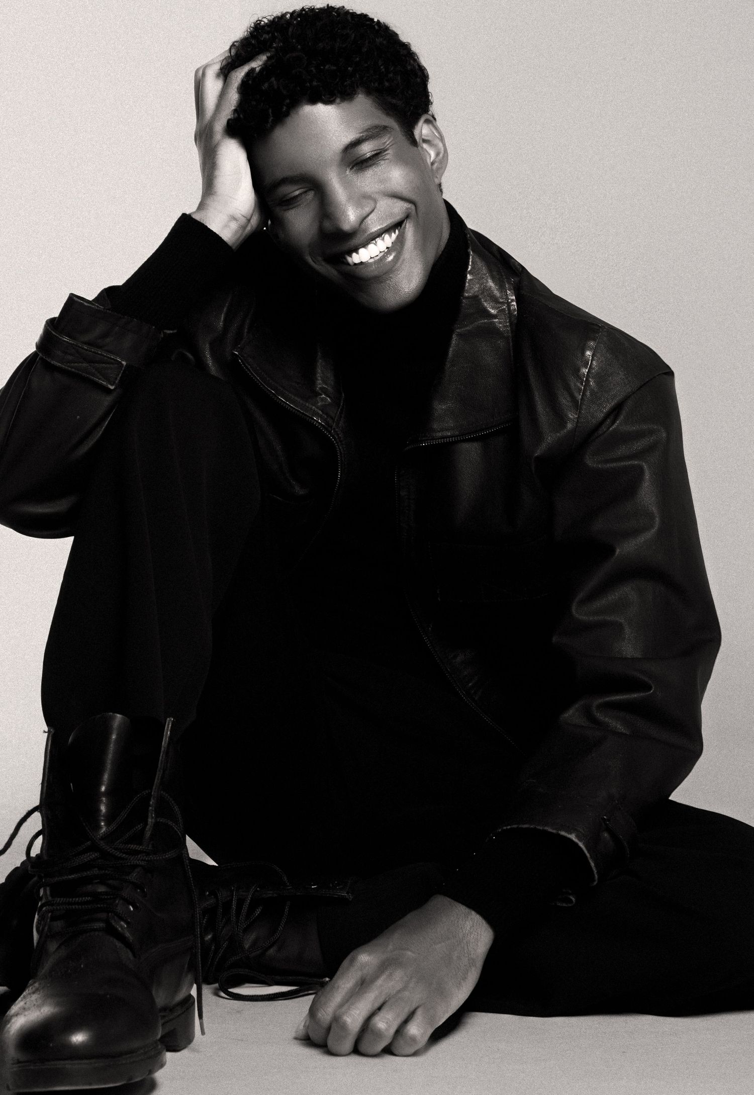 Smiling young man with curly hair sits laughing in a black leather jacket and lace-up boots in a black and white photo.