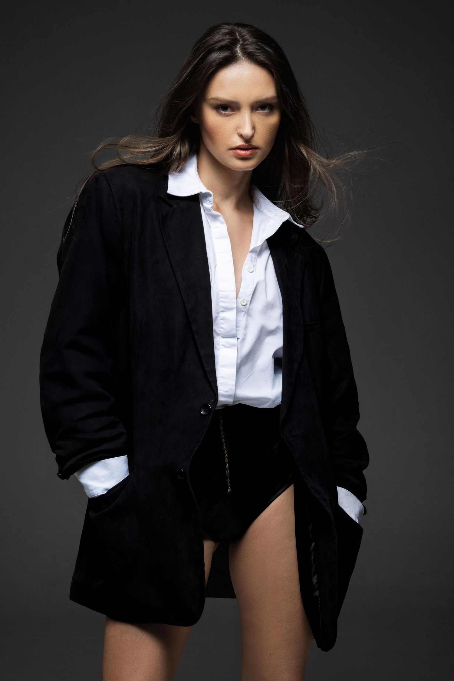 Model in an oversized black blazer and white shirt with hair in motion against a dark studio backdrop.