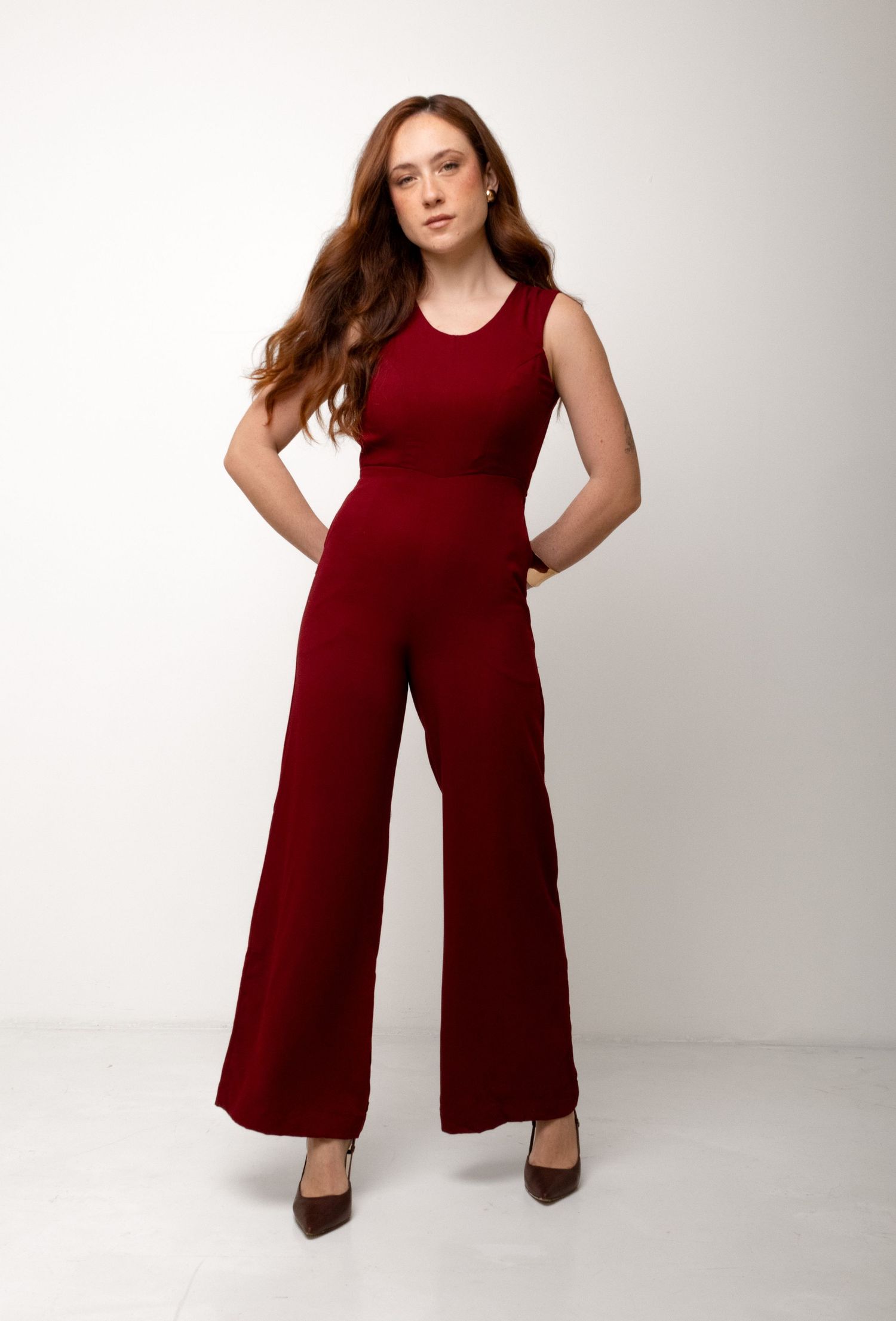 Woman with long auburn hair stands with hands on hips in a burgundy sleeveless wide-leg jumpsuit and dark heels.