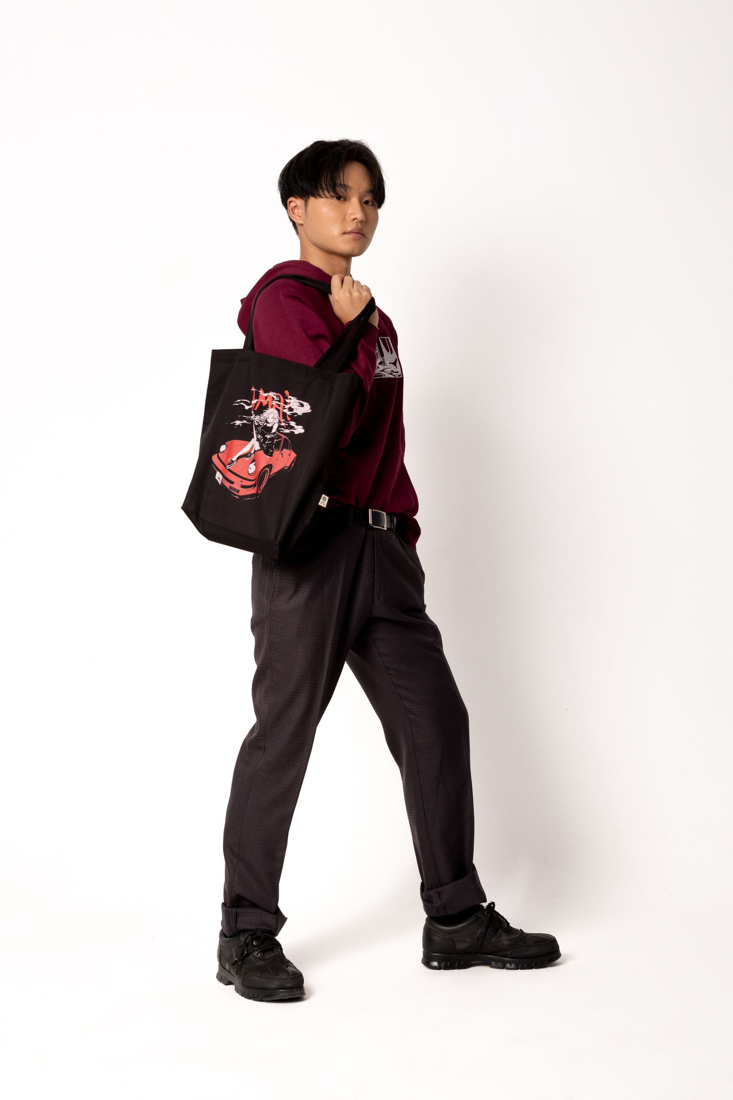 Man in a burgundy hoodie and gray trousers poses mid-stride with a black tote bag with a red car graphic slung over his shoulder.