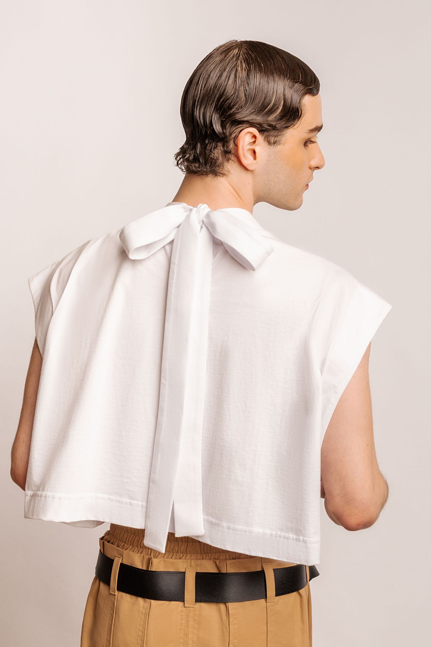 Close-up back view of a white sleeveless cropped top with a draped bow at the neck, worn with a black belt and tan trousers.