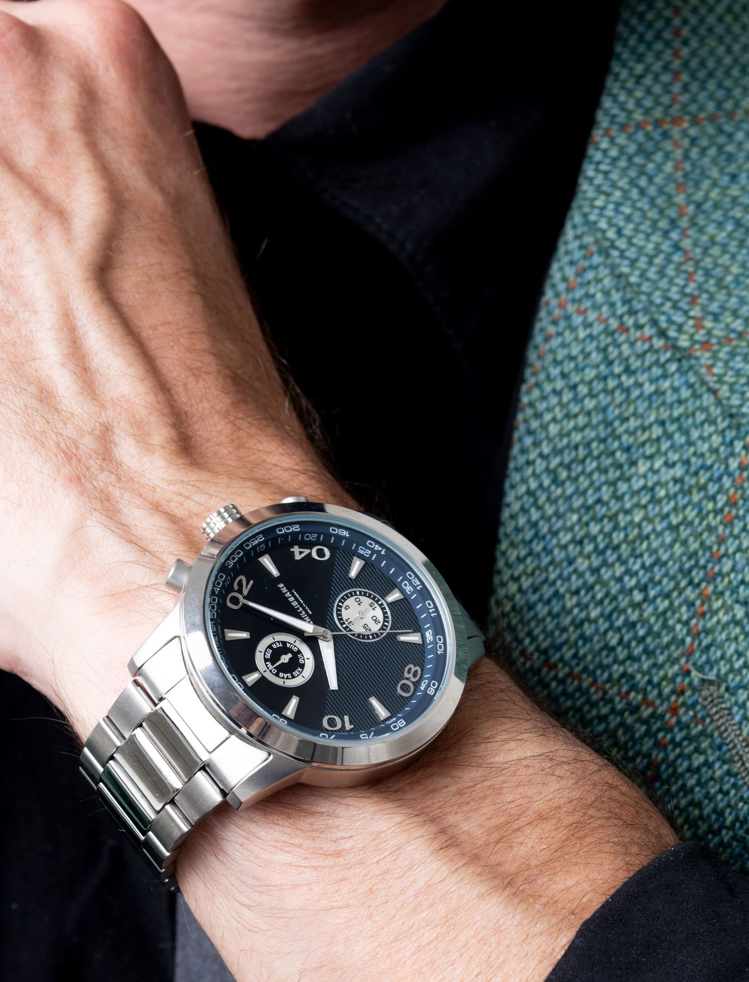 Close-up of a wrist wearing a silver stainless steel watch with a black dial, beside a green tweed jacket sleeve.