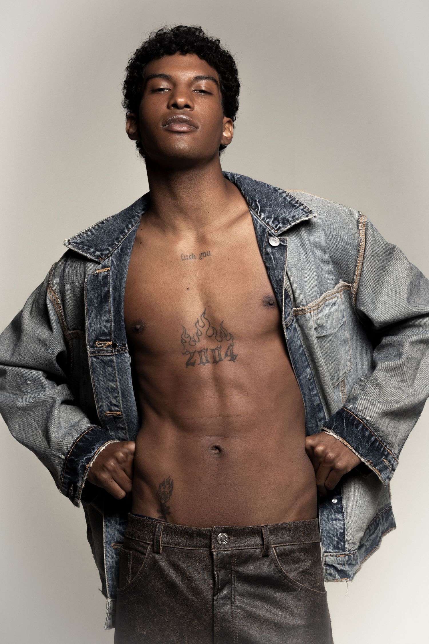 Young man with curly hair wears an open denim jacket and brown leather pants, showing chest tattoos with hands on his hips.
