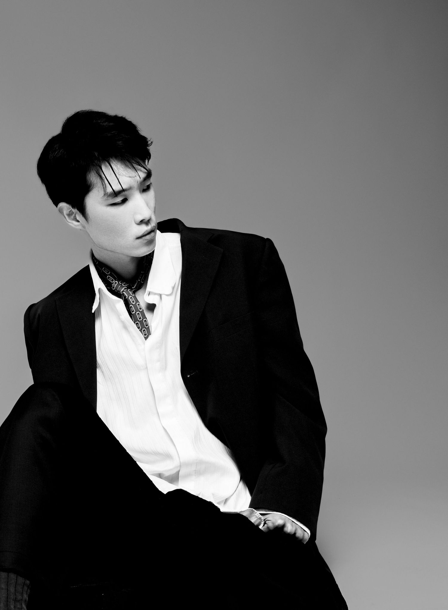 Black and white fashion portrait of a seated man in a dark blazer, white shirt and patterned scarf, looking away.