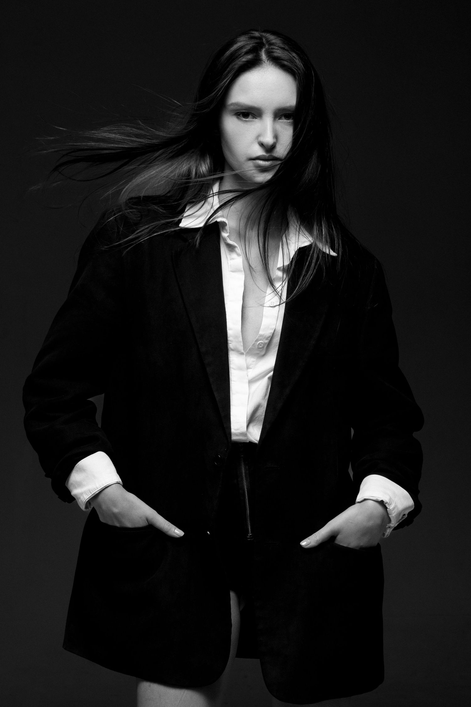 Black and white portrait of a model in an oversized blazer and open white shirt, hands in pockets, hair flowing.
