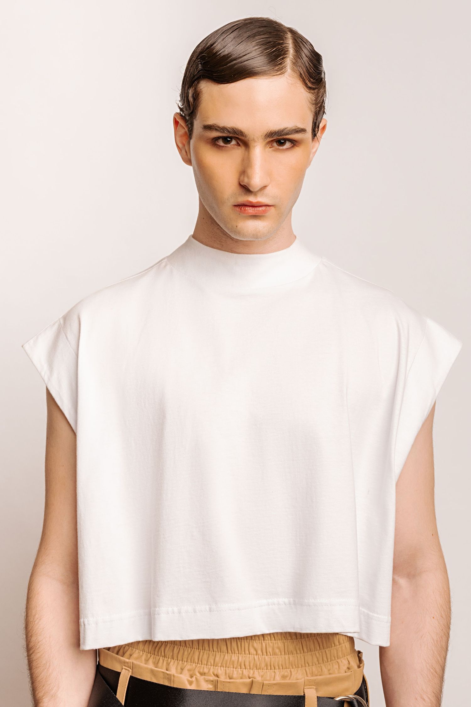Front portrait of a model in a white mock-neck sleeveless cropped top over a tan elastic waistband with a sleek side-parted hairstyle.