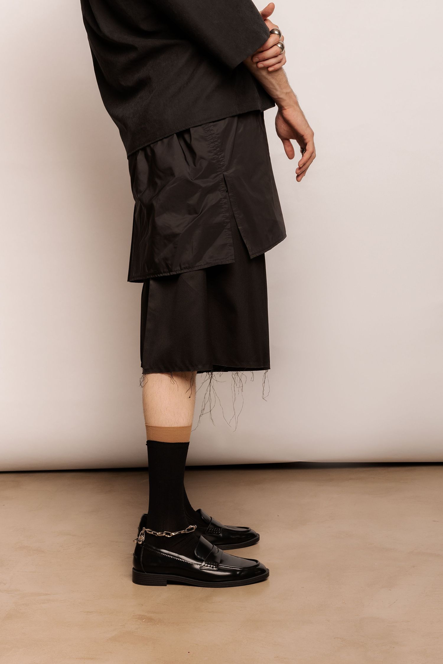 Side view of layered black shorts with raw frayed hems, black socks and black loafers with a silver chain detail.