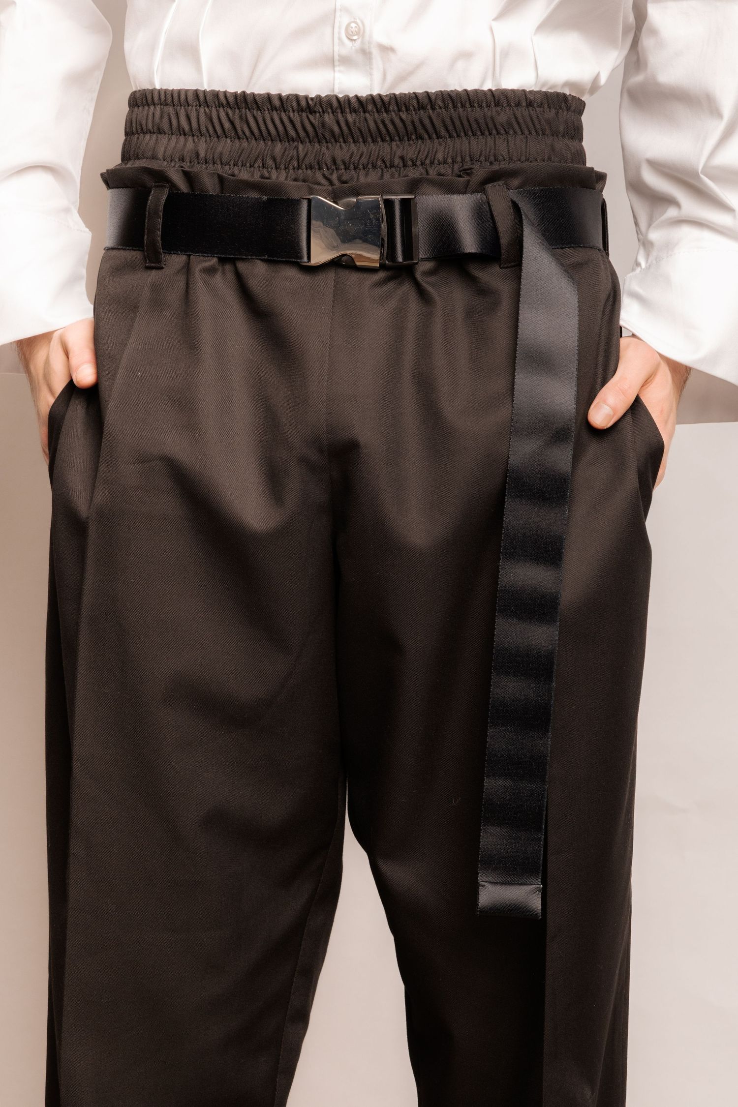Close-up of black elastic-waist trousers with a black buckled webbing belt, worn under an untucked white shirt with hands in pockets.