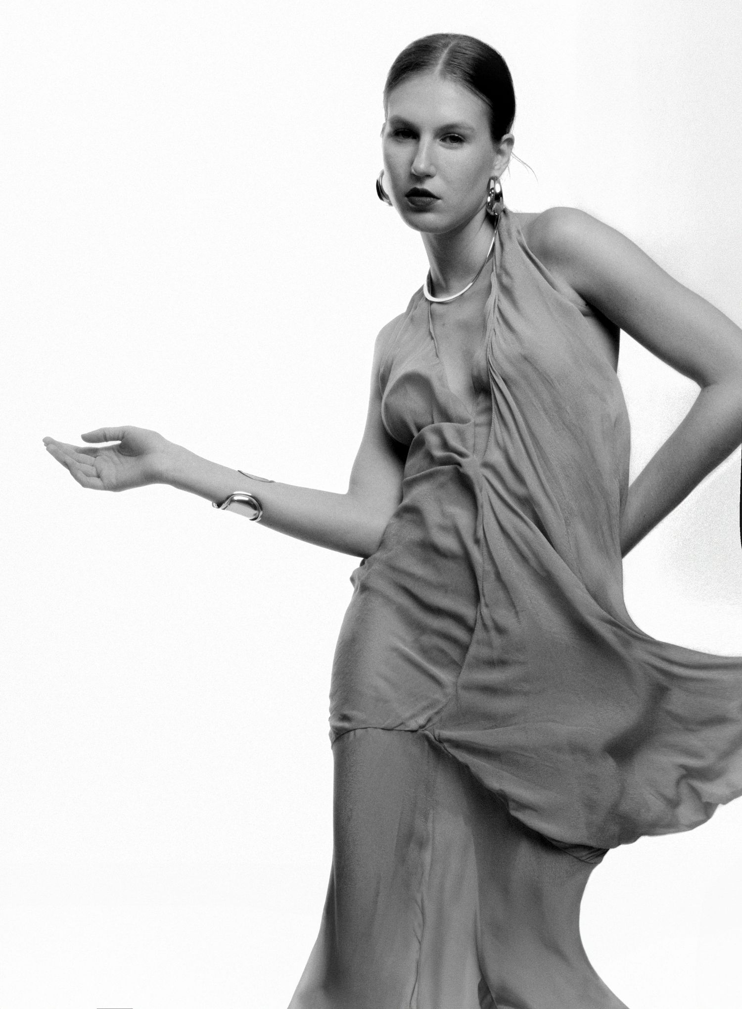 Black and white fashion portrait of a model in a flowing draped halter dress with hoop earrings and a bracelet, one arm extended.