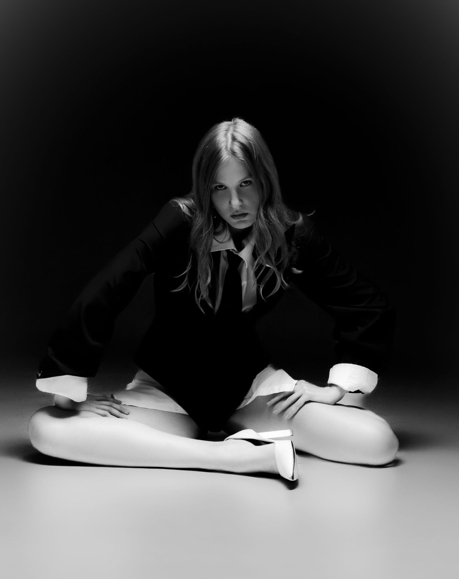 Moody black and white fashion photo of a model sitting on the floor in a dark blazer, tie, white tights and heels.