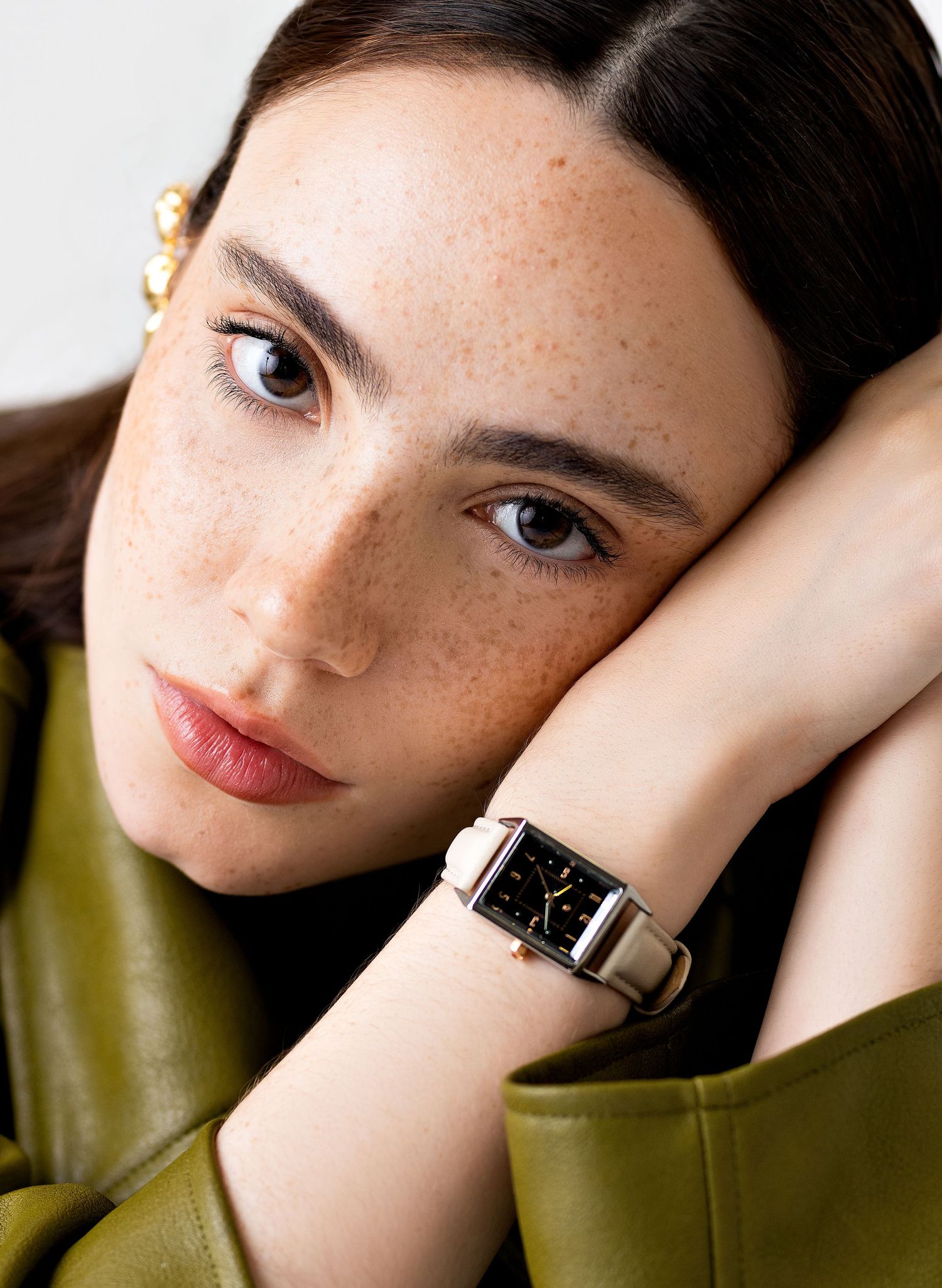 Freckled woman in an olive green leather jacket rests her head on her arm, wearing a rectangular watch with a white leather strap.