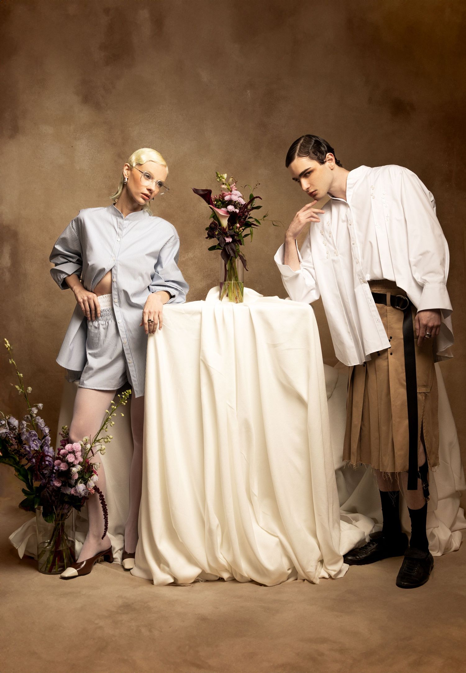 Two models in light oversized shirts and shorts pose beside a draped white table with flower arrangements against a textured brown backdrop.