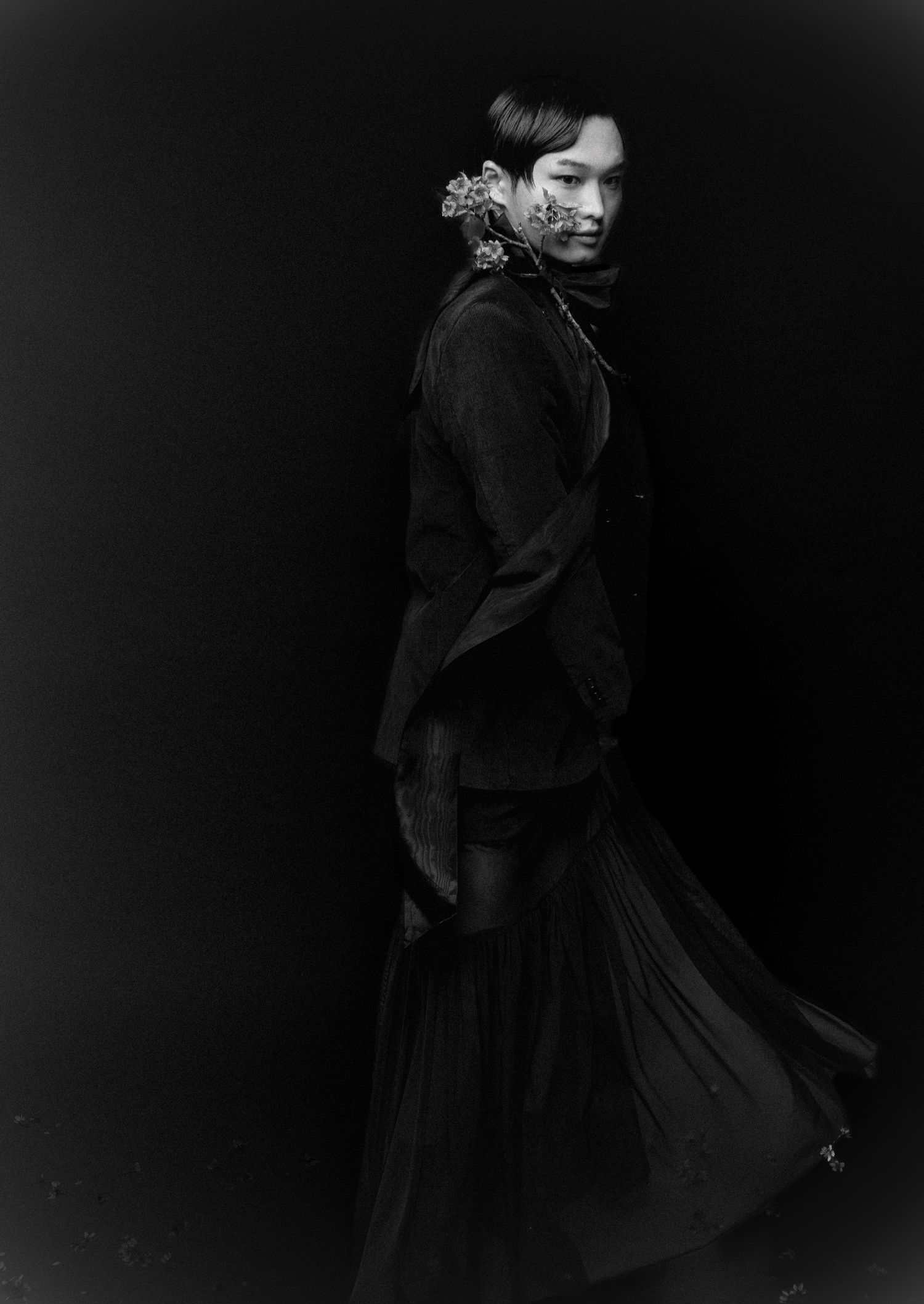 Moody black and white fashion image of a model in a dark layered pleated skirt and jacket, with flowers at the shoulder.