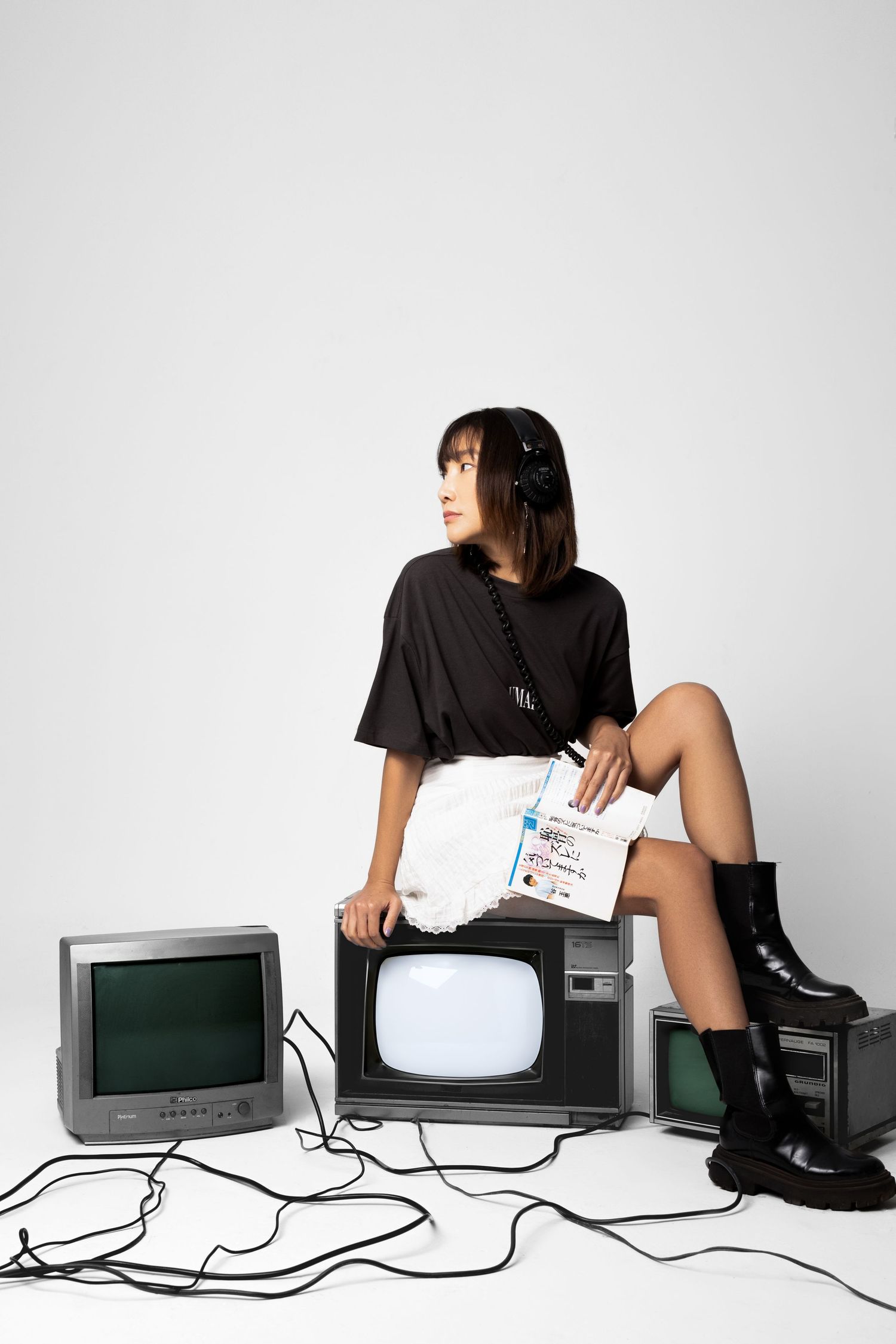 Woman in black tee, white shorts, headphones and boots sits on vintage television sets holding a booklet against a white backdrop.