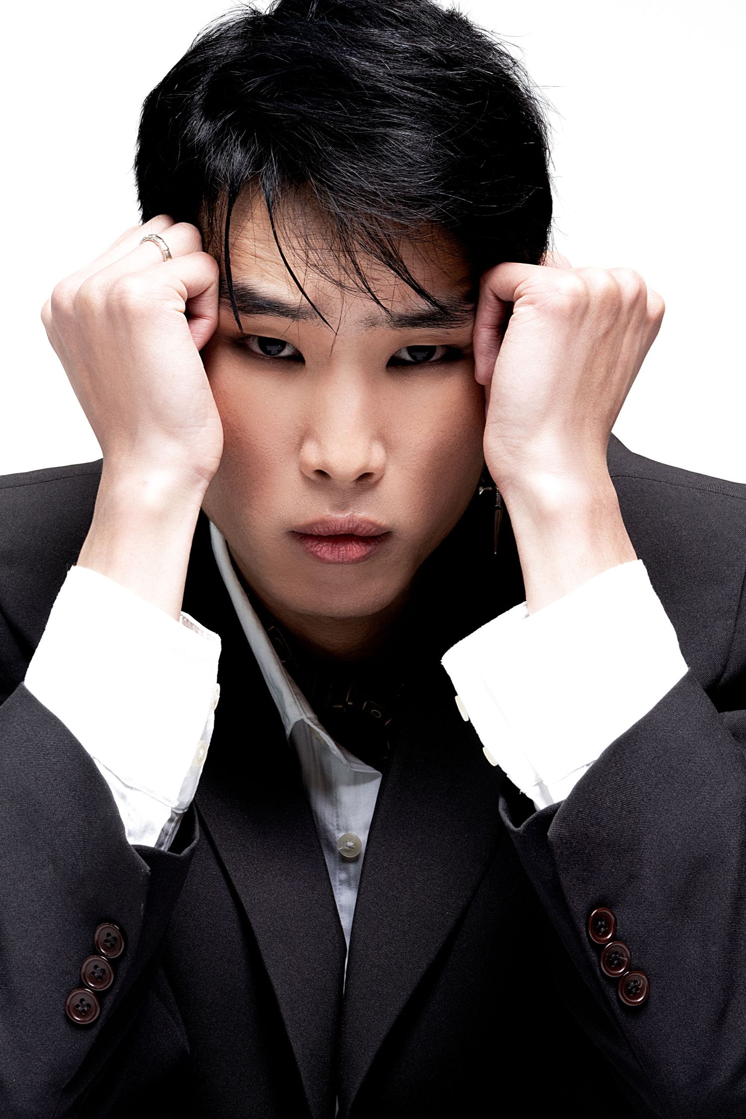 Young man in a dark suit presses both hands against his head, looking intensely at the camera in a studio portrait.