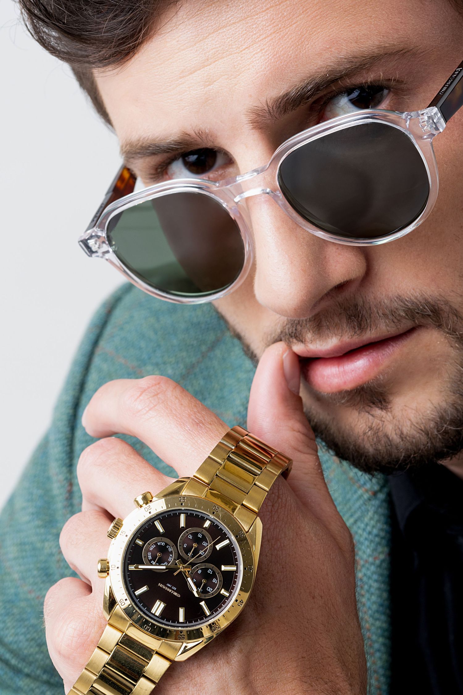 Close-up of a man in clear-framed sunglasses holding up his wrist to show a gold chronograph watch with a black dial.