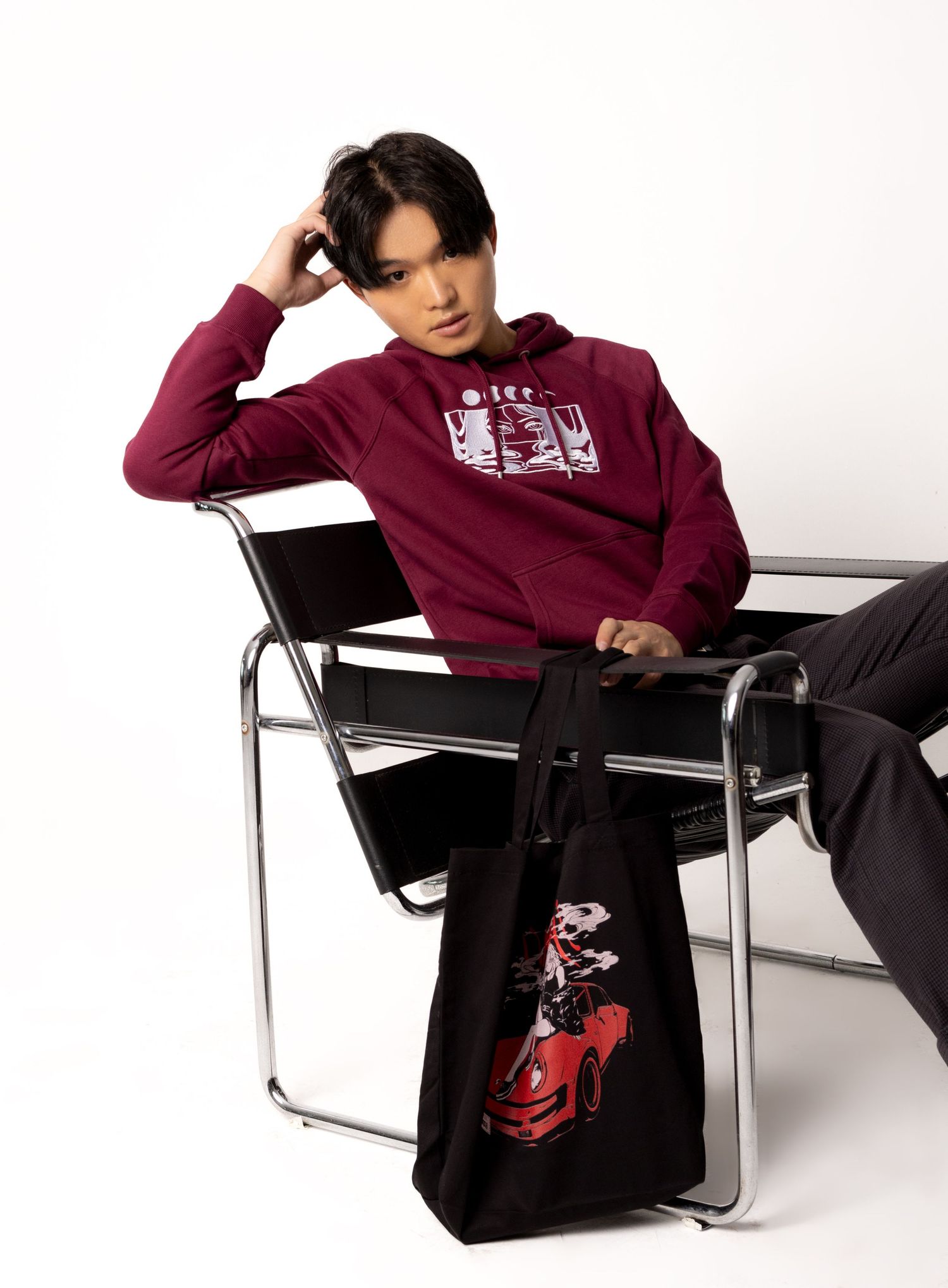 Man in a burgundy graphic hoodie and dark trousers sits in a chrome Wassily-style chair with a black tote featuring a red car print.