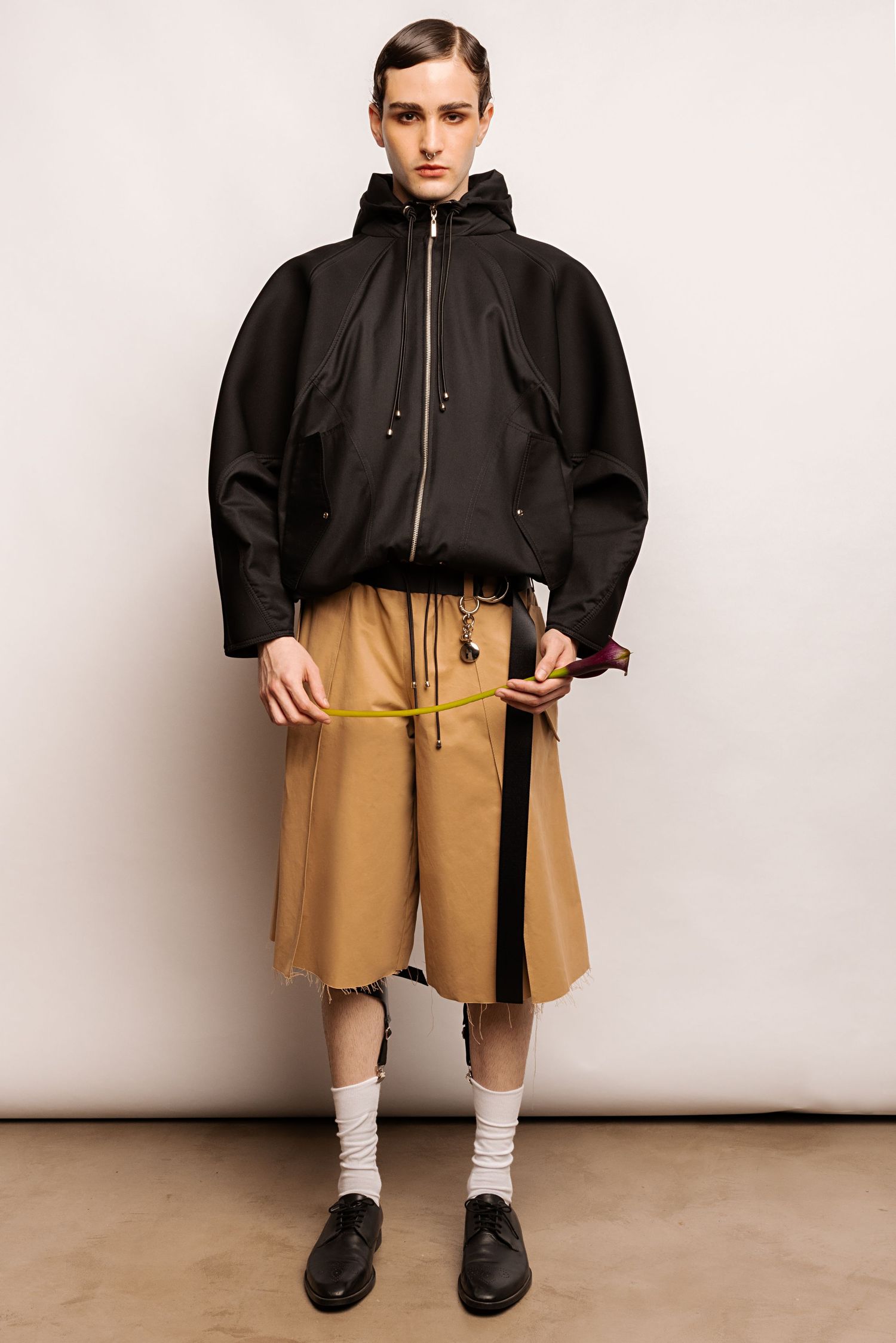 Model in an oversized black hooded jacket, khaki bermuda shorts, white socks and black lace-up shoes stands against a plain backdrop.