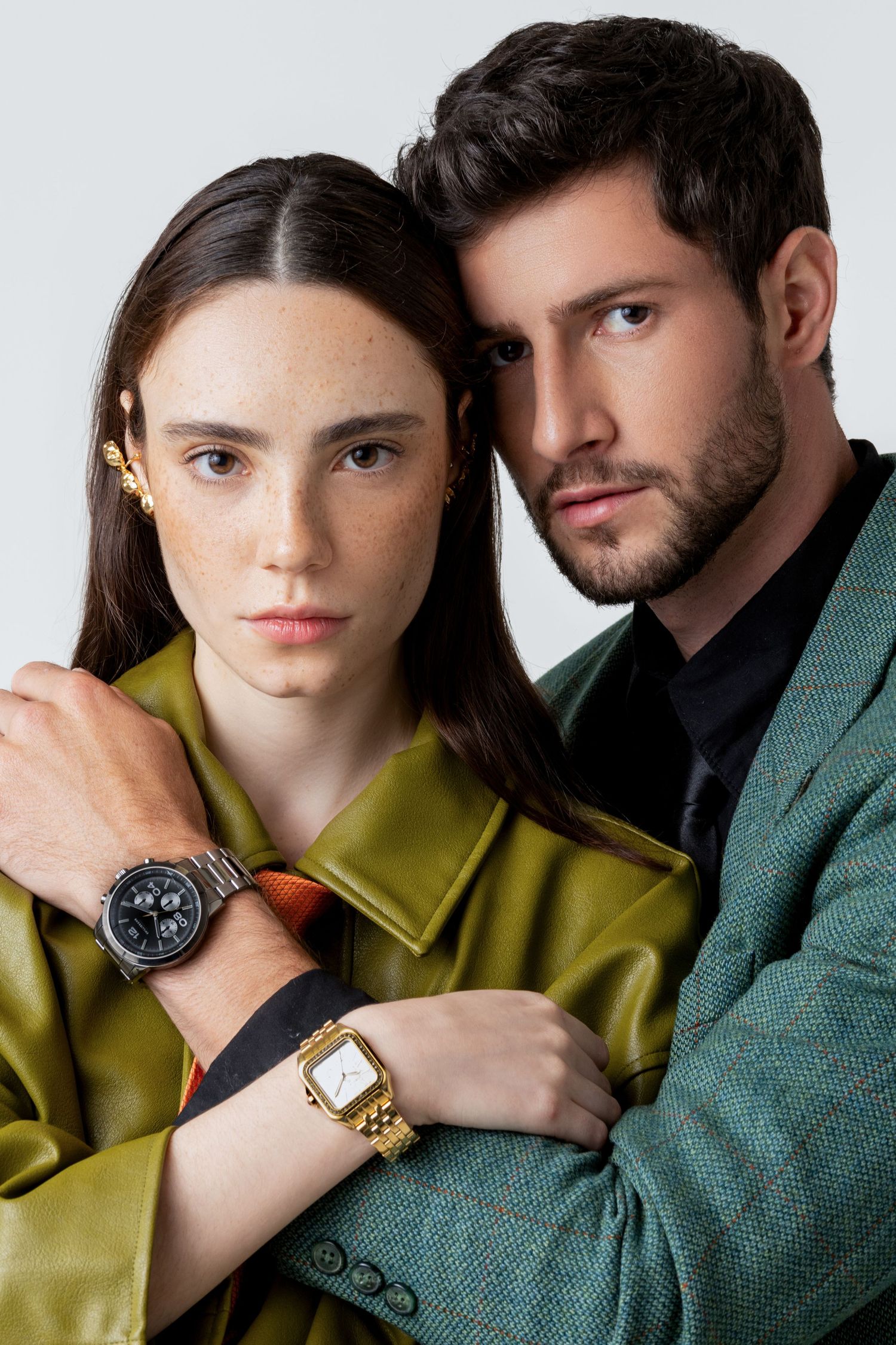 Woman in an olive jacket leans against a man in a green blazer, her arm showing a gold square watch and a black chronograph.