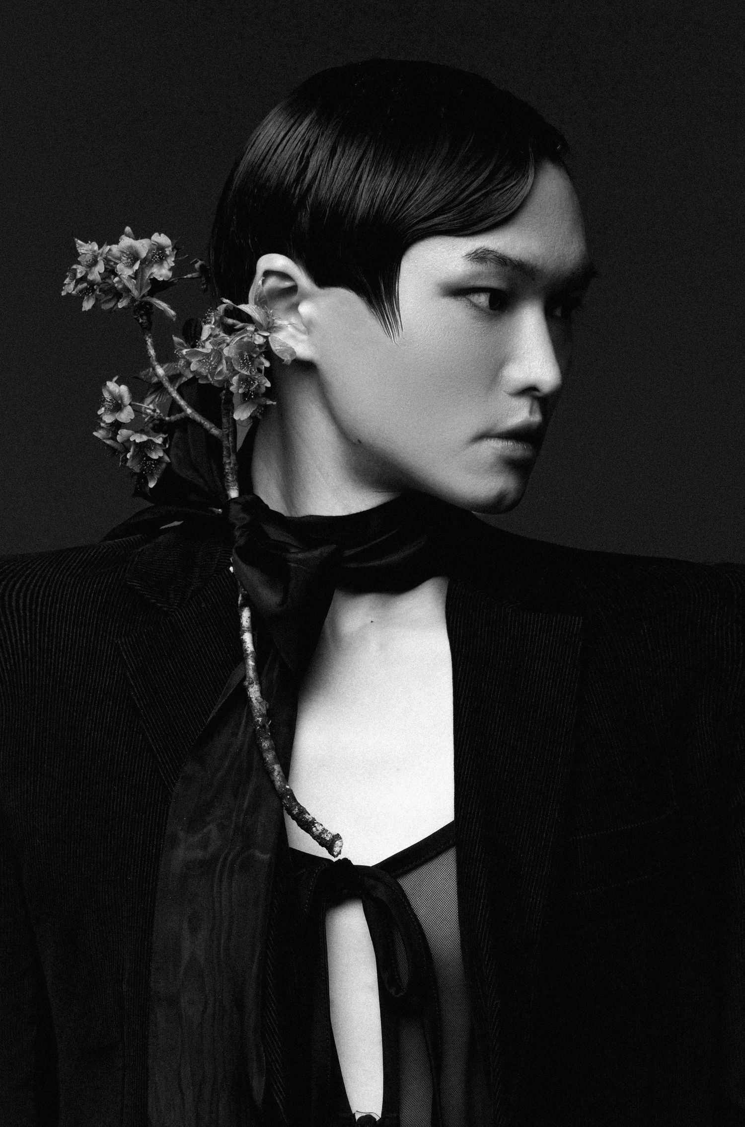 Black and white fashion portrait of a model in profile with a slicked hairstyle and a flower branch tucked behind the ear.