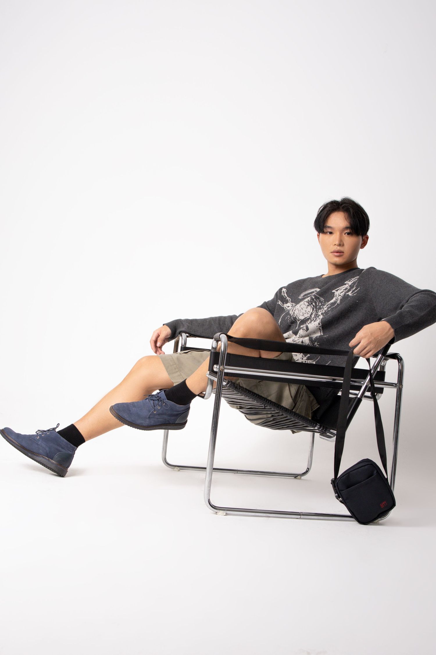 Model in a gray graphic sweater and navy shoes lounges in a chrome-framed chair with a black bag at his side.