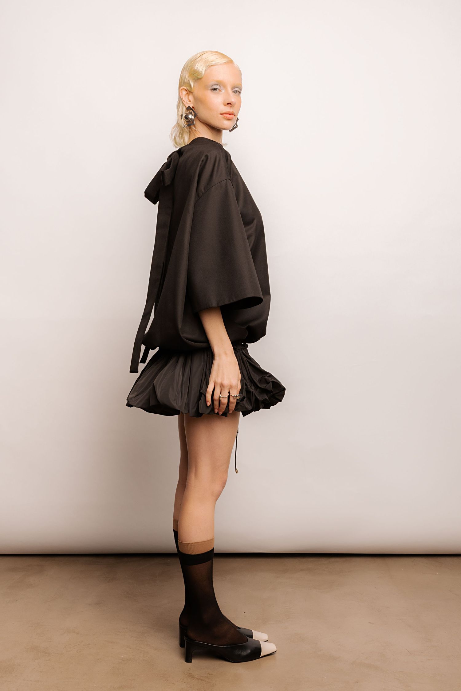 Side profile of a model in a black wide-sleeve top and puffed ruffle mini skirt with sheer black socks and two-tone heels.