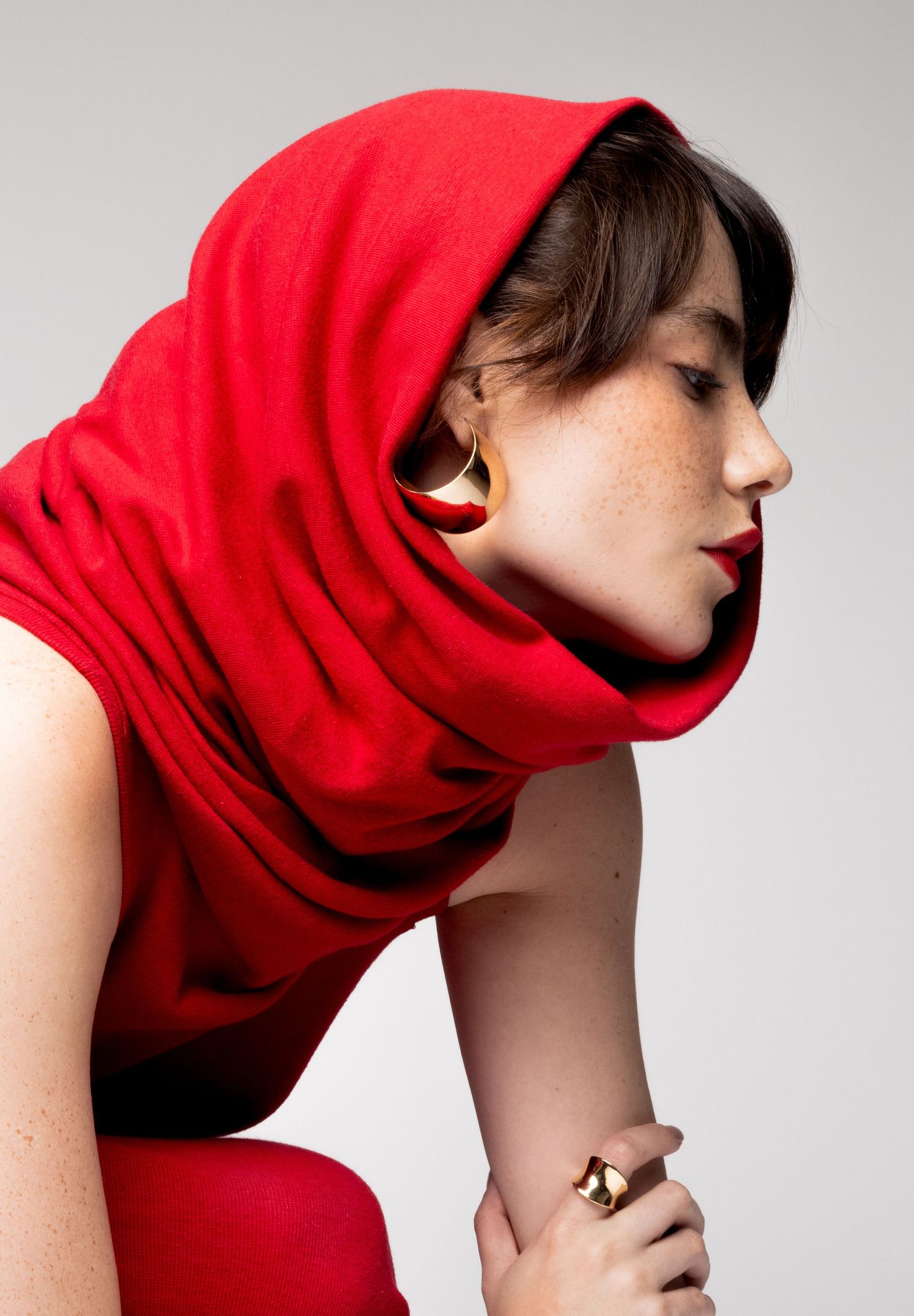 Side profile of a freckled woman in a red hooded cowl top with a gold sculptural earring and ring, leaning forward.