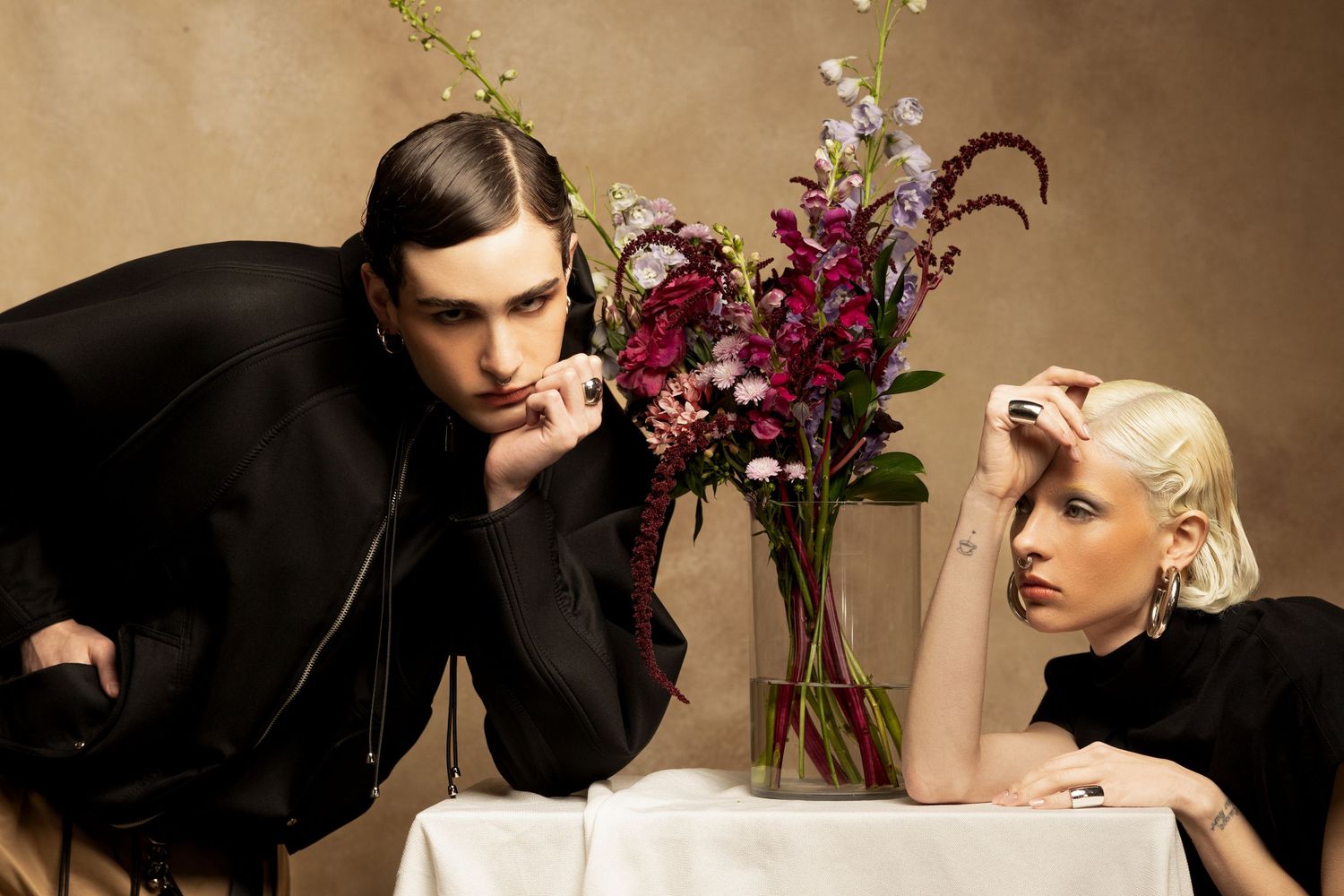 Two models in black fashion pieces pose at a white table with a vase of pink and burgundy flowers in a moody editorial scene.