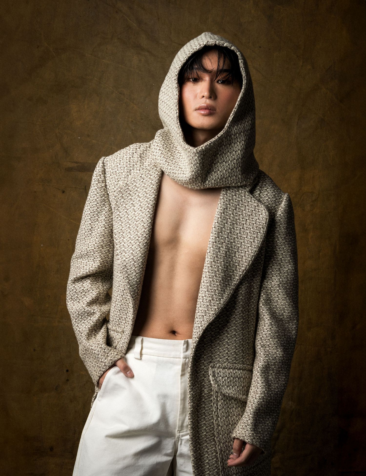 Model in a hooded tweed coat worn open over a bare chest and white trousers, posed against a moody golden-brown backdrop.