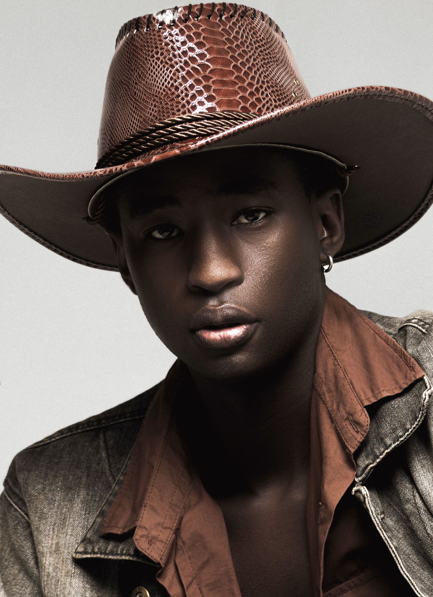 Close-up portrait of a male model in a glossy brown snakeskin-style cowboy hat and open rust-colored shirt with a gray denim jacket.