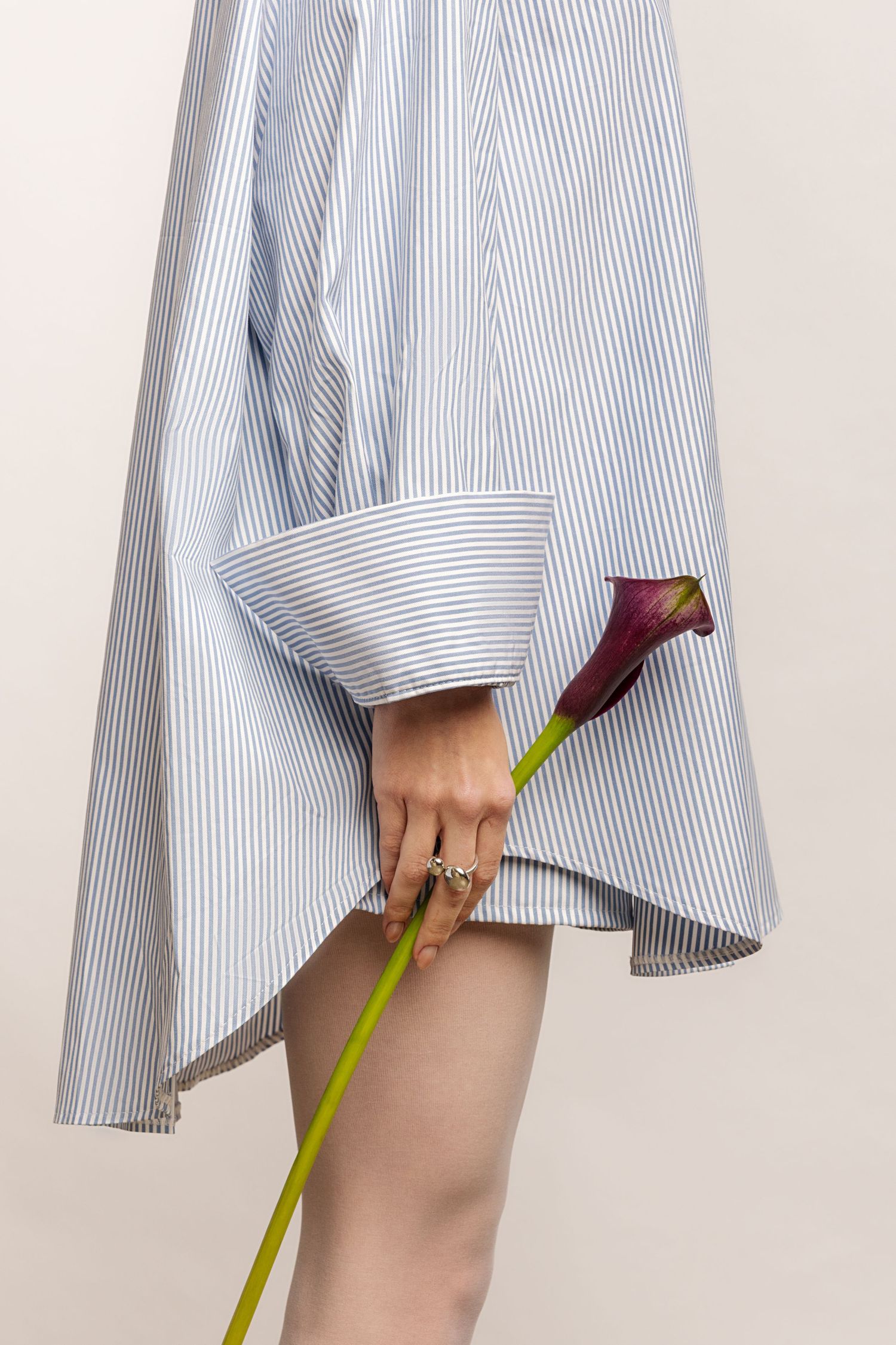 Close-up of a hand with silver rings holding a purple calla lily beside an oversized blue striped shirt with a wide cuff.