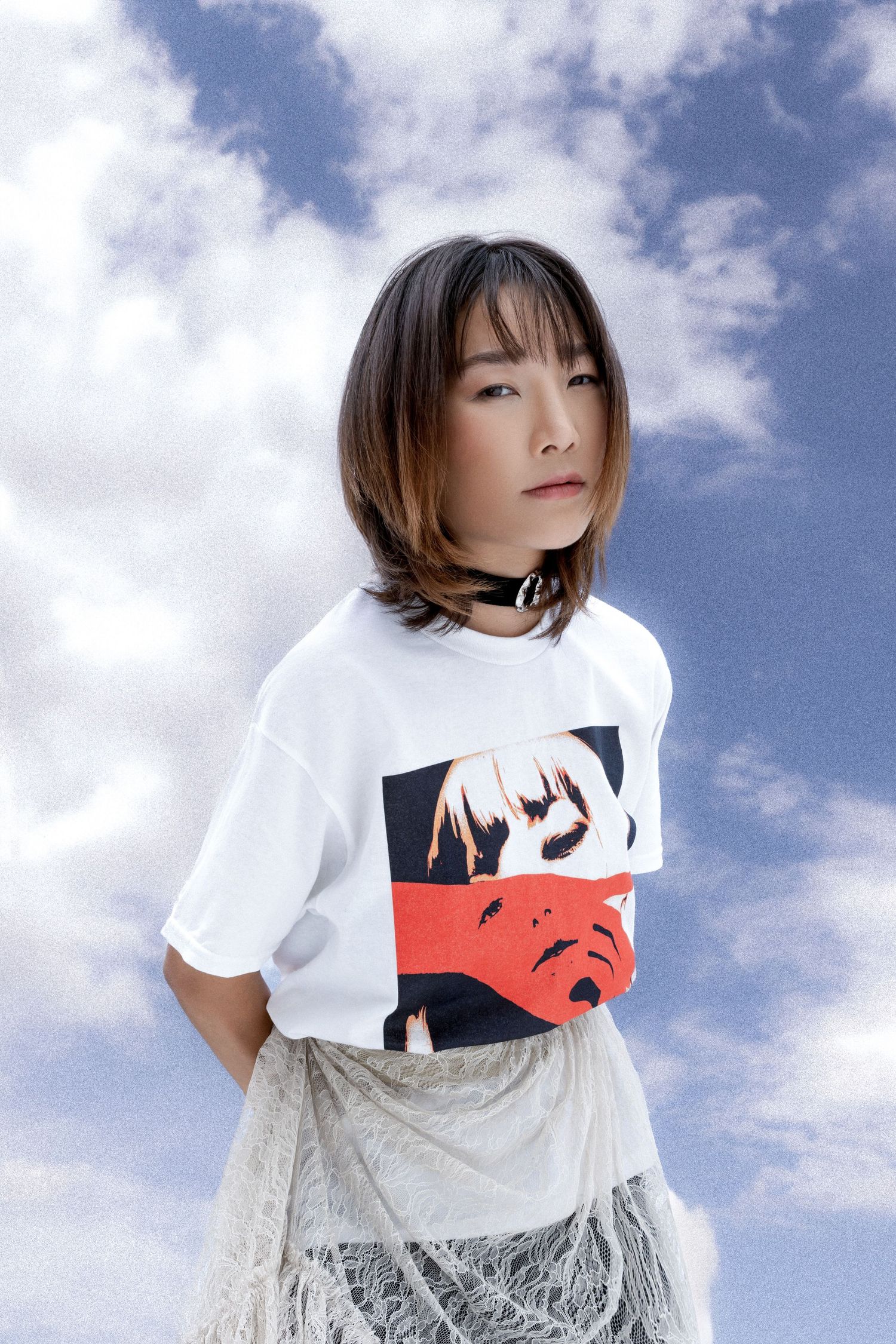 Woman with a layered bob and choker wears a white graphic tee over a lace skirt, posed against a bright blue cloudy sky.