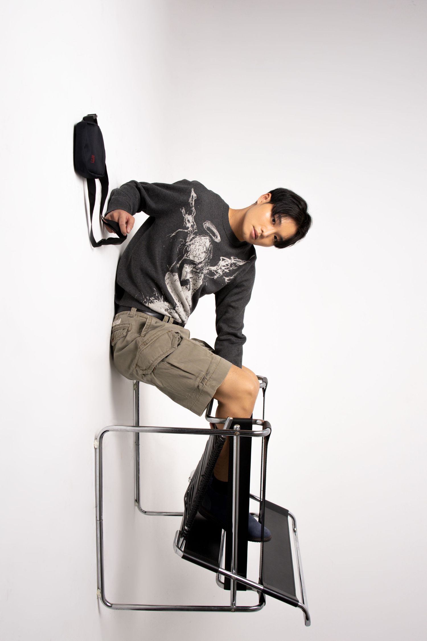 Model in a graphic sweater and cargo shorts poses on a chrome chair tilted against a white wall with a black bag.