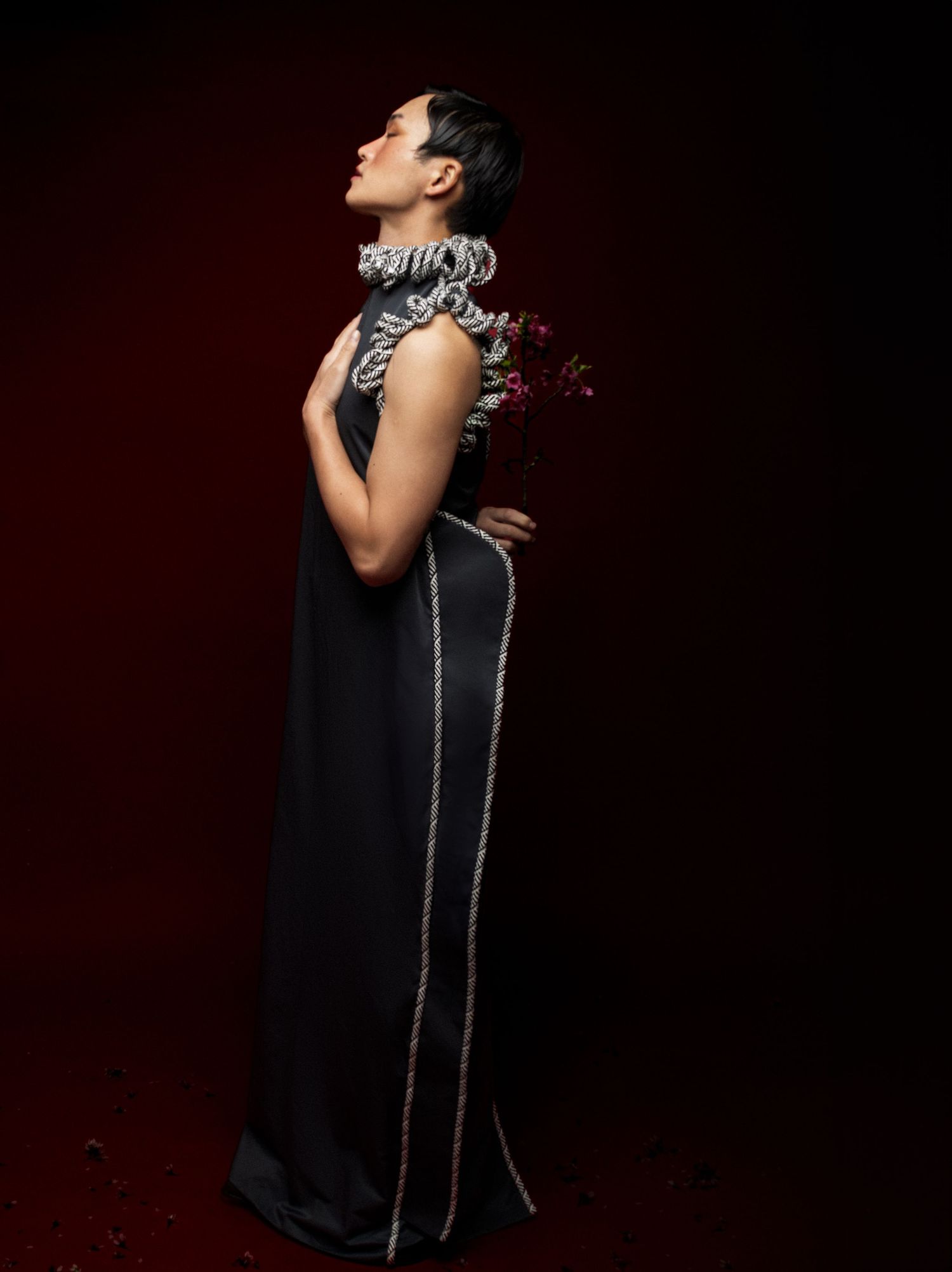 Model gazes upward in a long black gown with silver trim and ruffled collar, standing against a dark crimson studio backdrop.