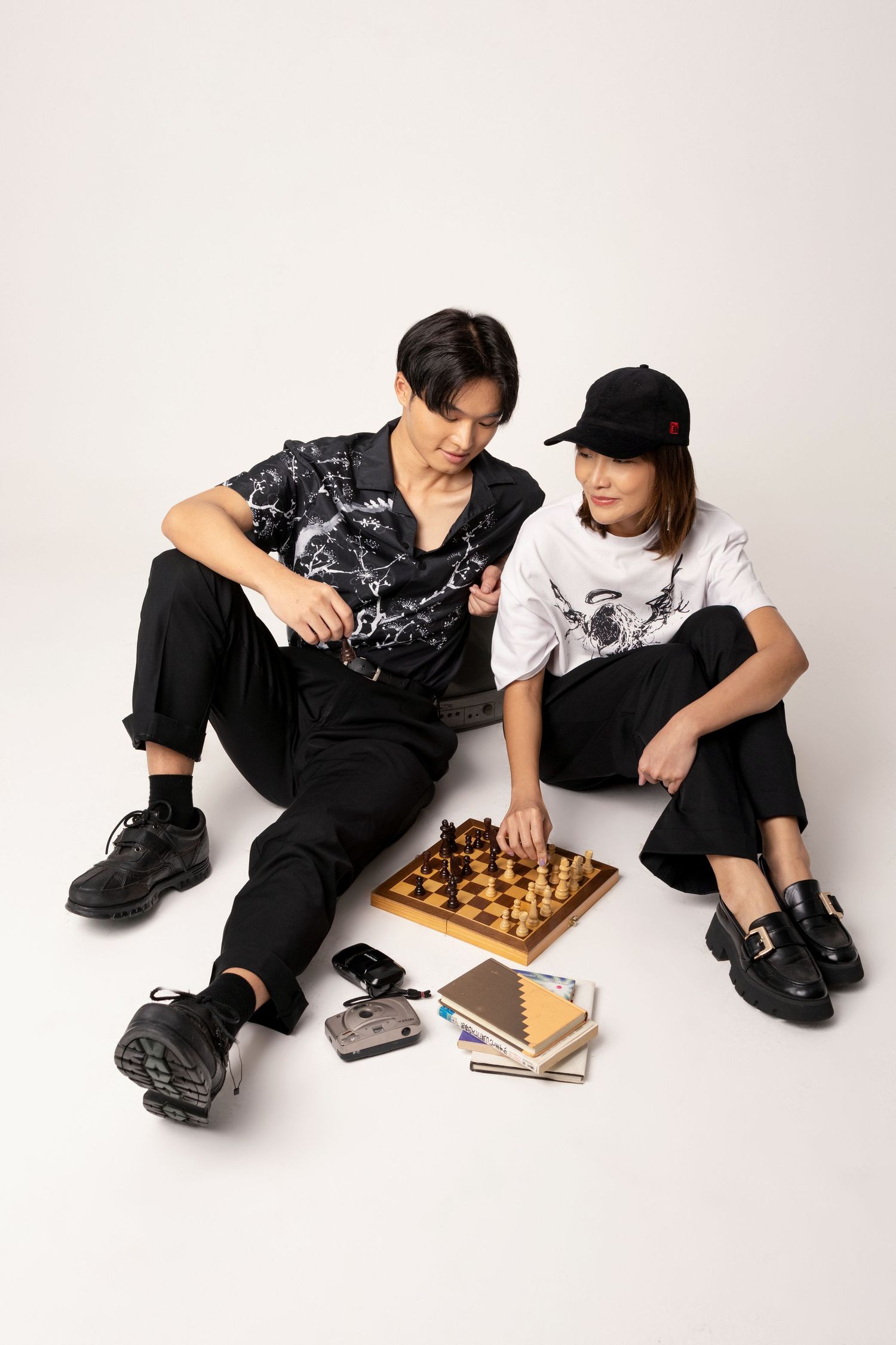A man and a woman in a cap sit on a white floor playing chess, surrounded by books and a compact camera, wearing black streetwear.