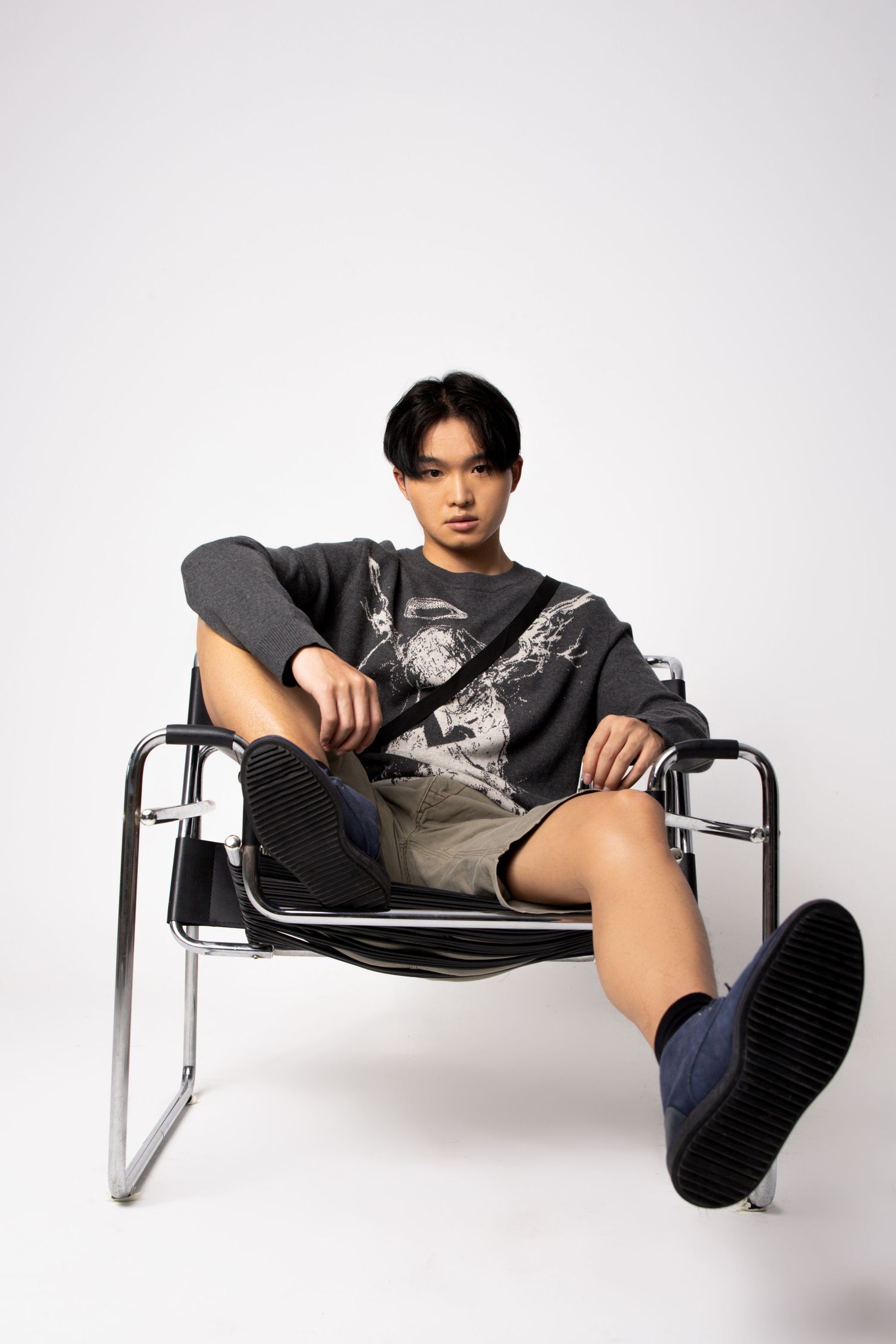 Young man in a gray graphic sweater, khaki shorts and navy shoes sits in a tubular steel chair in a studio.