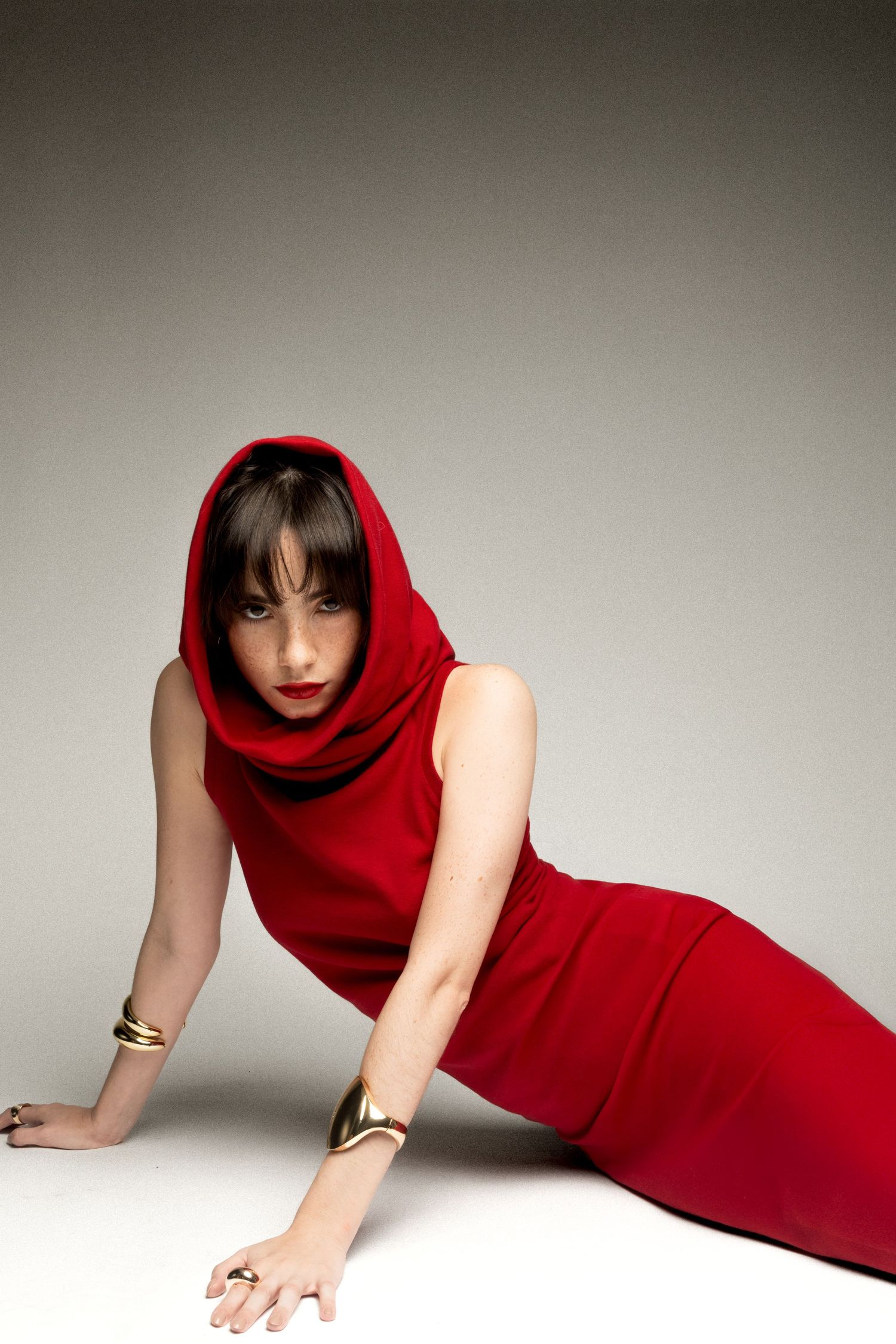 Woman in a red hooded sleeveless gown reclines on a white studio floor, wearing gold cuff bracelets and rings.