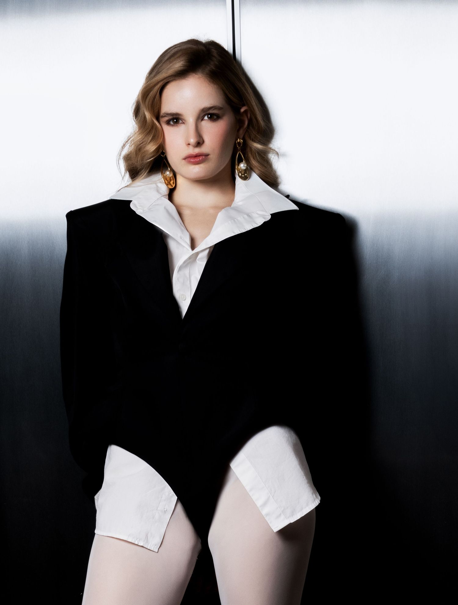 Model with wavy hair and gold earrings wearing an oversized black blazer over a white shirt and white tights against a metallic wall.