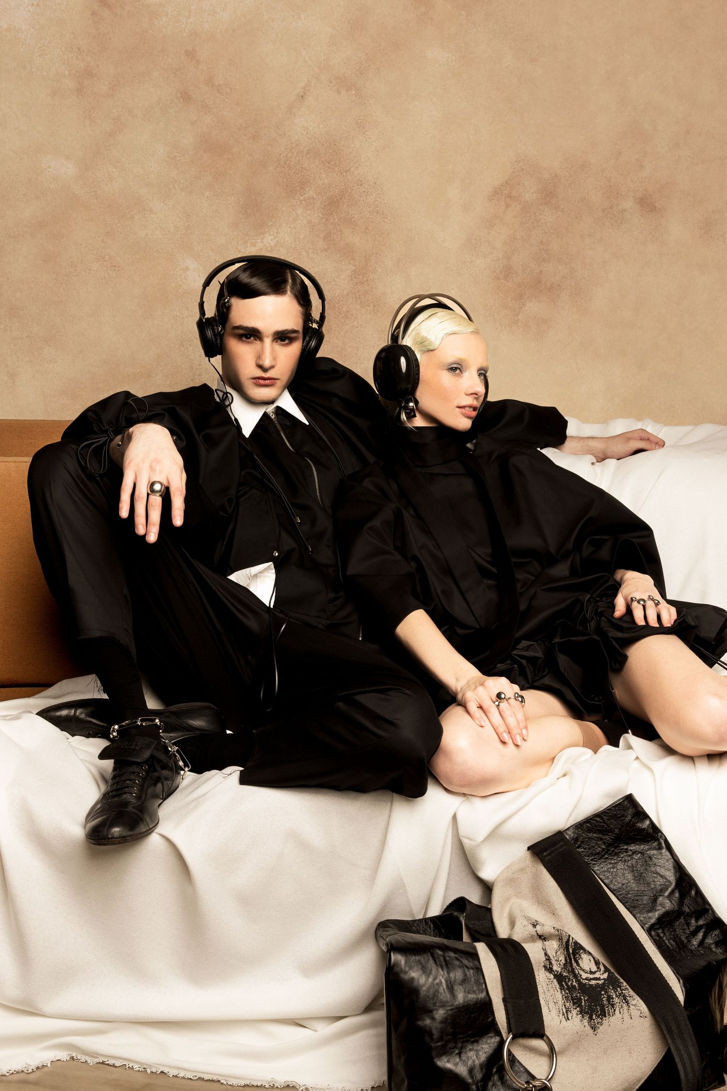 Two models in black layered fashion and headphones lounge on a sofa draped in white fabric beside a distressed black leather bag.