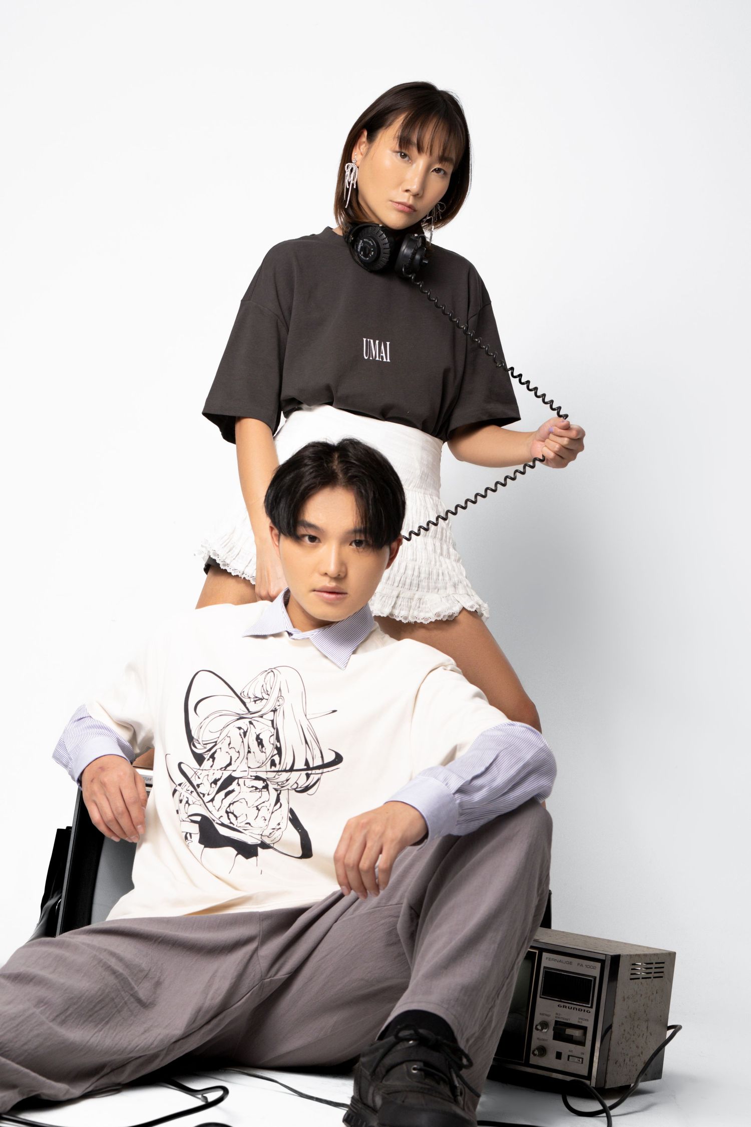 A woman in a gray tee and ruffled white skirt holds headphones above a seated man in a graphic shirt in a bright studio.