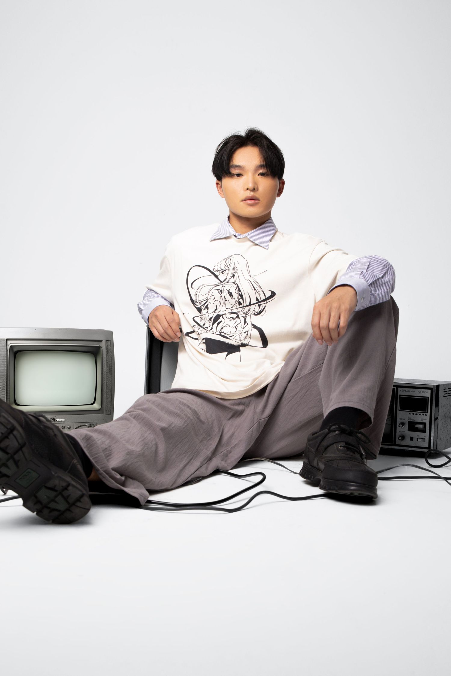 A model in a graphic vest, layered shirt and chunky black shoes sits on the floor among vintage TVs and cables.
