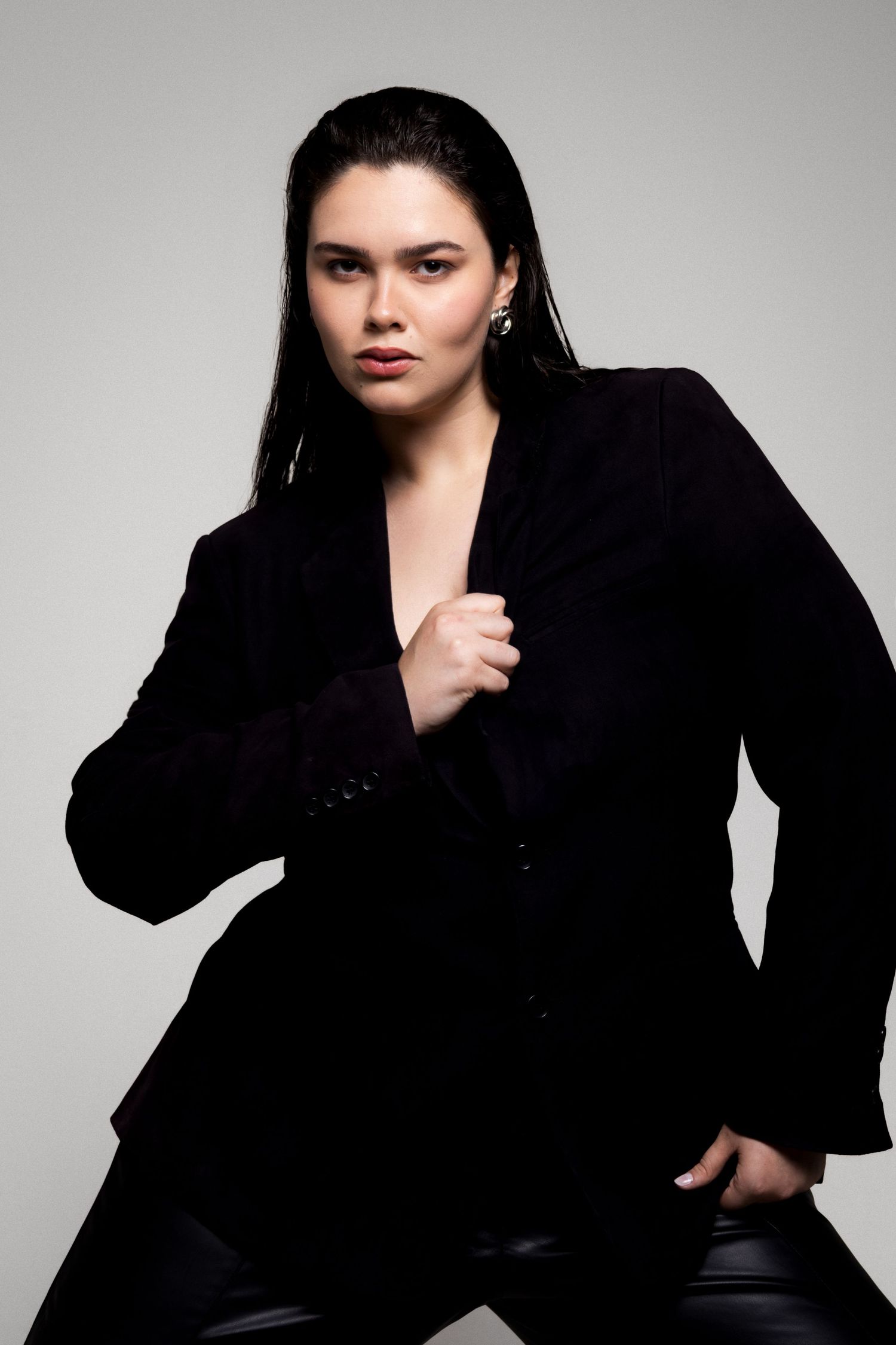Model with slicked-back dark hair wears a black blazer over leather pants and holds the lapel while posing against a gray background.