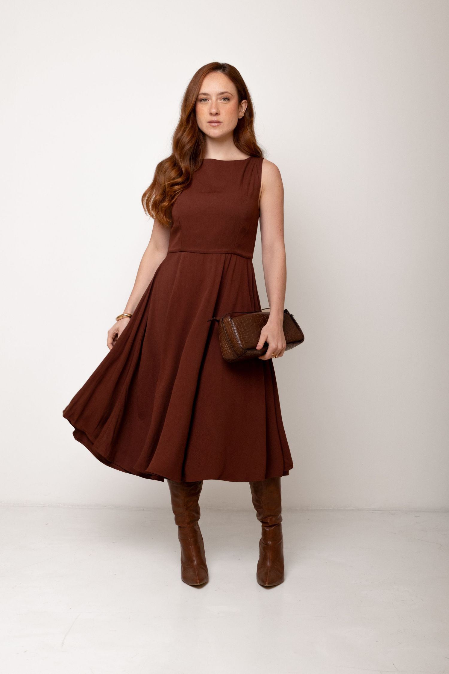 Model wearing a sleeveless chocolate brown midi dress with a full skirt, holding a brown clutch and wearing knee-high brown boots.