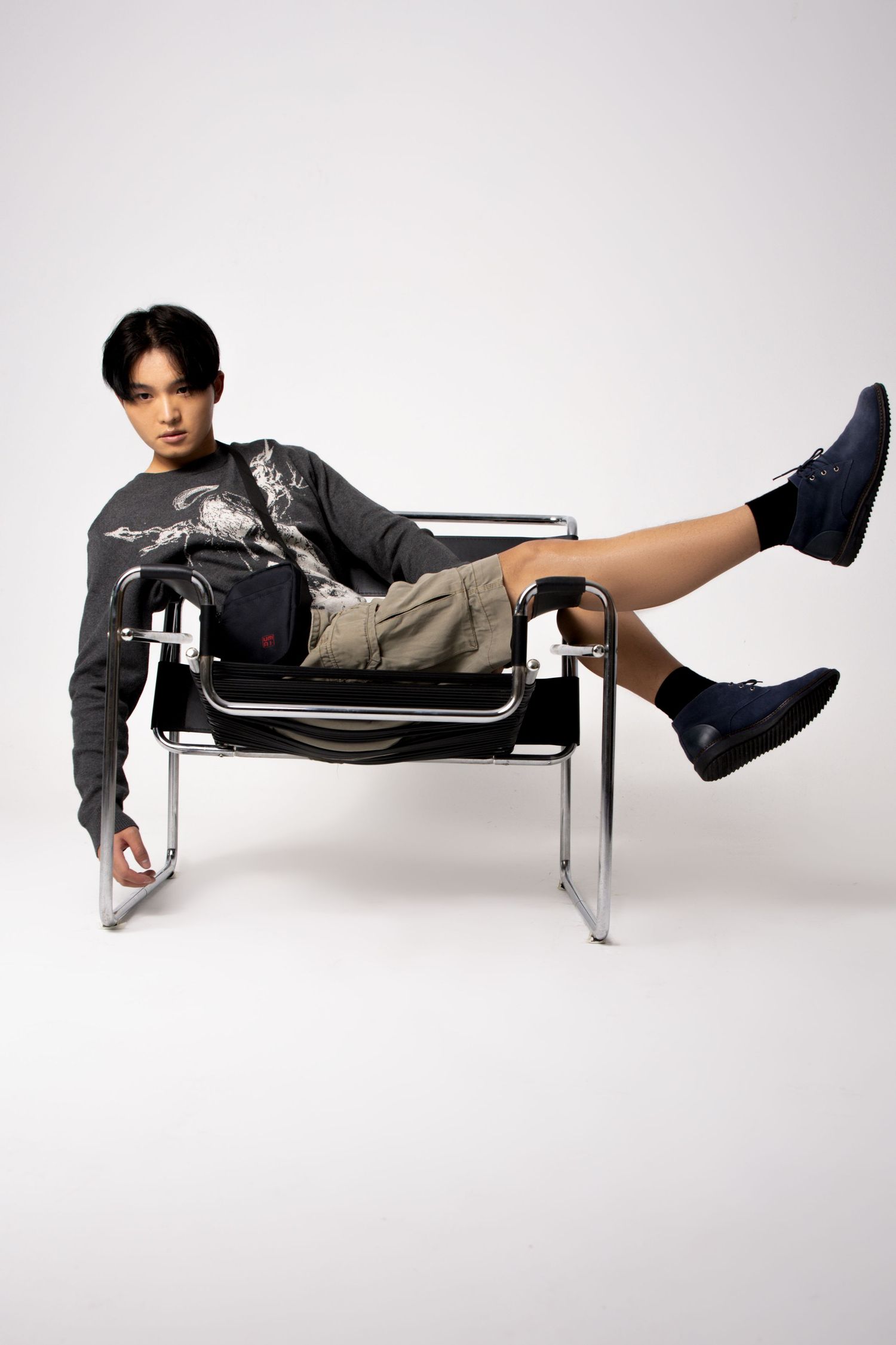 Model in a graphic gray sweater and cargo shorts reclines in a steel-framed chair with legs raised in a studio.