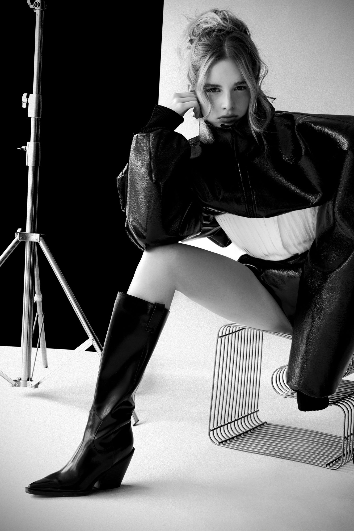 Black and white fashion shot of a model in a leather jacket and tall black boots seated on a wire chair in a photo studio.