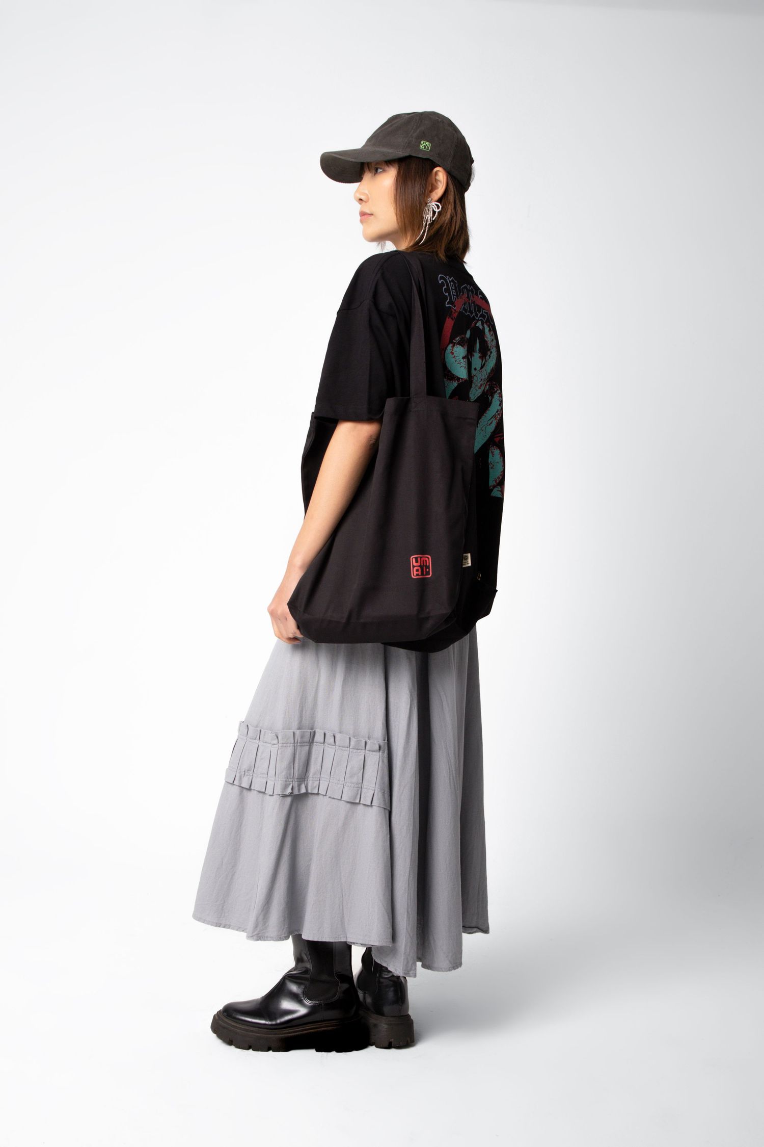 Woman in profile wearing a black tee with a back graphic, gray tiered skirt, cap and boots, carrying a black tote bag on her shoulder.