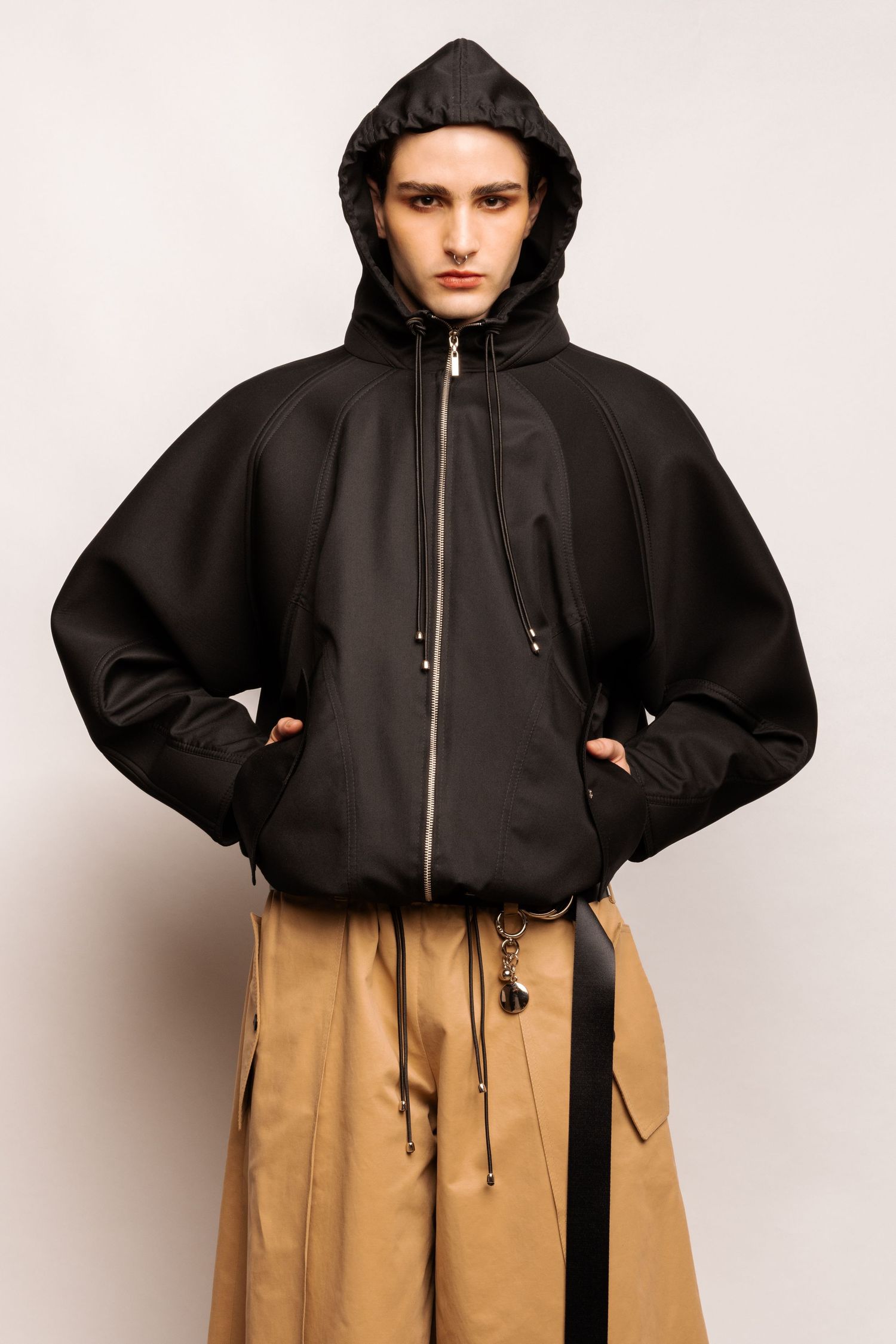 Close-up of a model wearing a black oversized hooded jacket with drawstrings and tan cargo trousers with a black belt and charm.