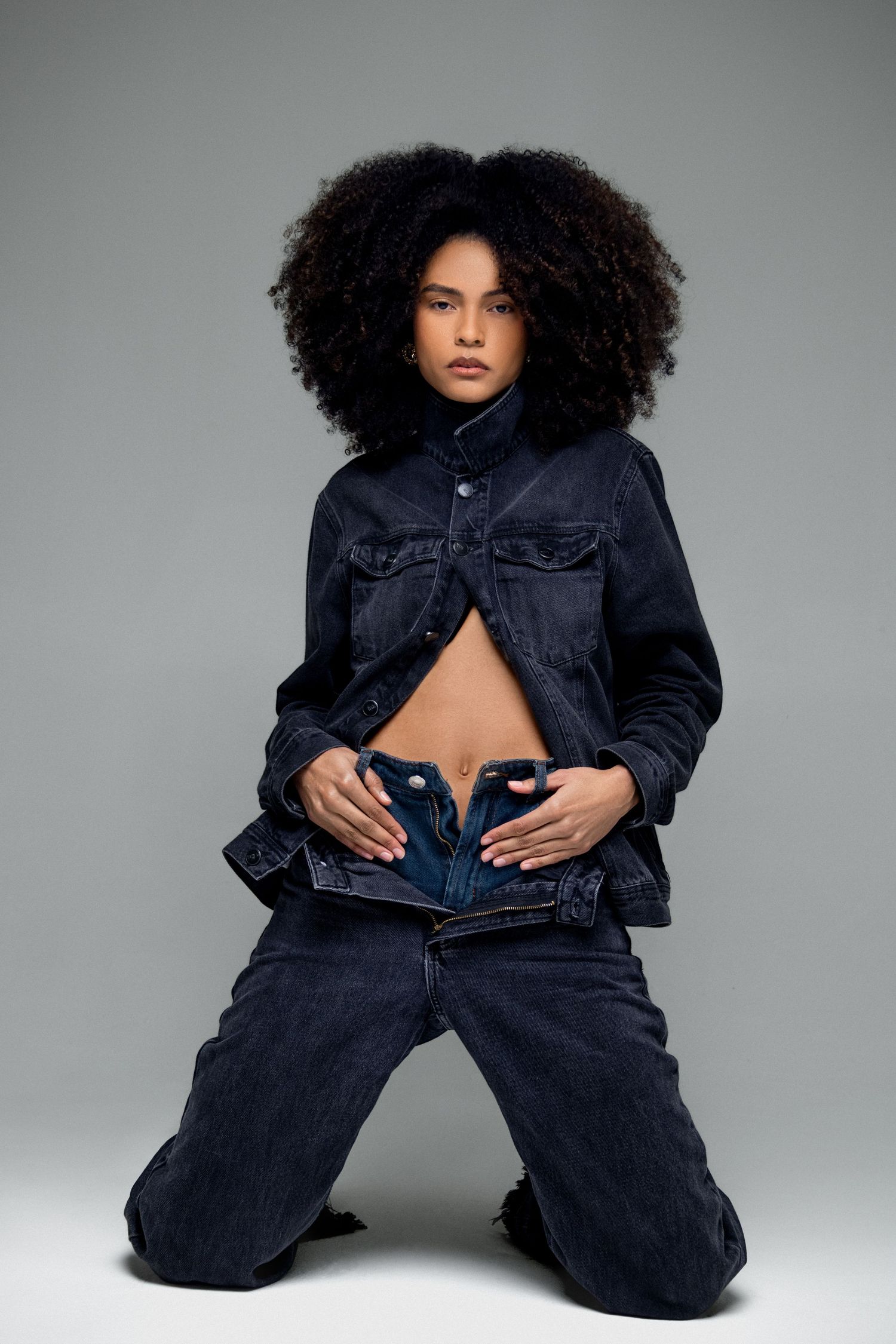 Model with voluminous natural curls kneels in a dark denim jacket and jeans, unbuttoned at the waist, against a gray studio backdrop.